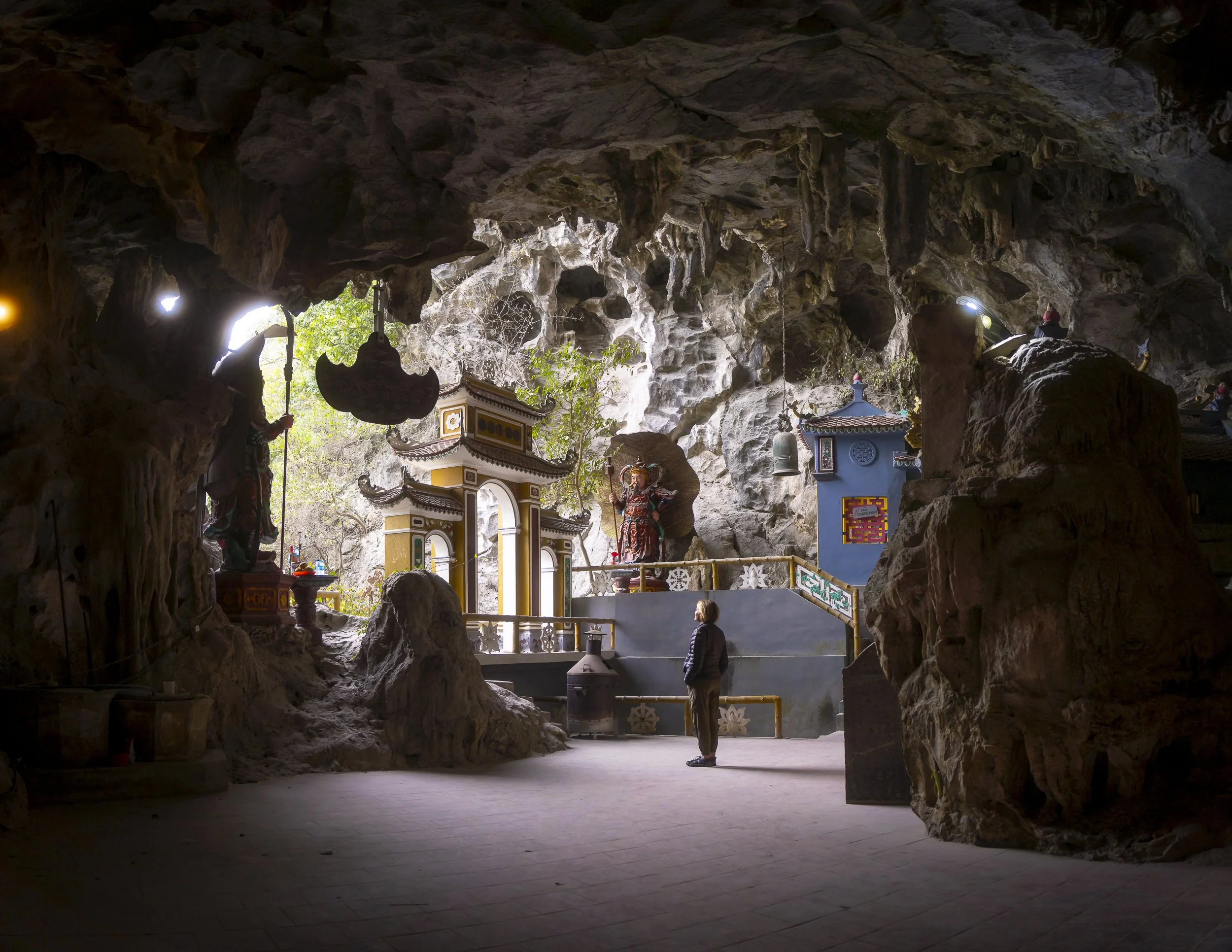 A person standing inside a cave temple with statues, ornate structures, and hanging bells, illuminated by natural light from outside.