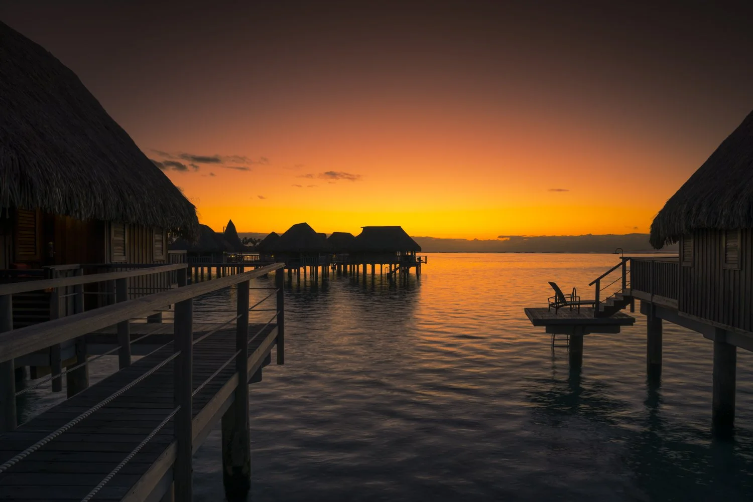 Overwater bungalows with thatched roofs at sunset, reflecting on calm water, with a colorful sky of orange and pink hues.