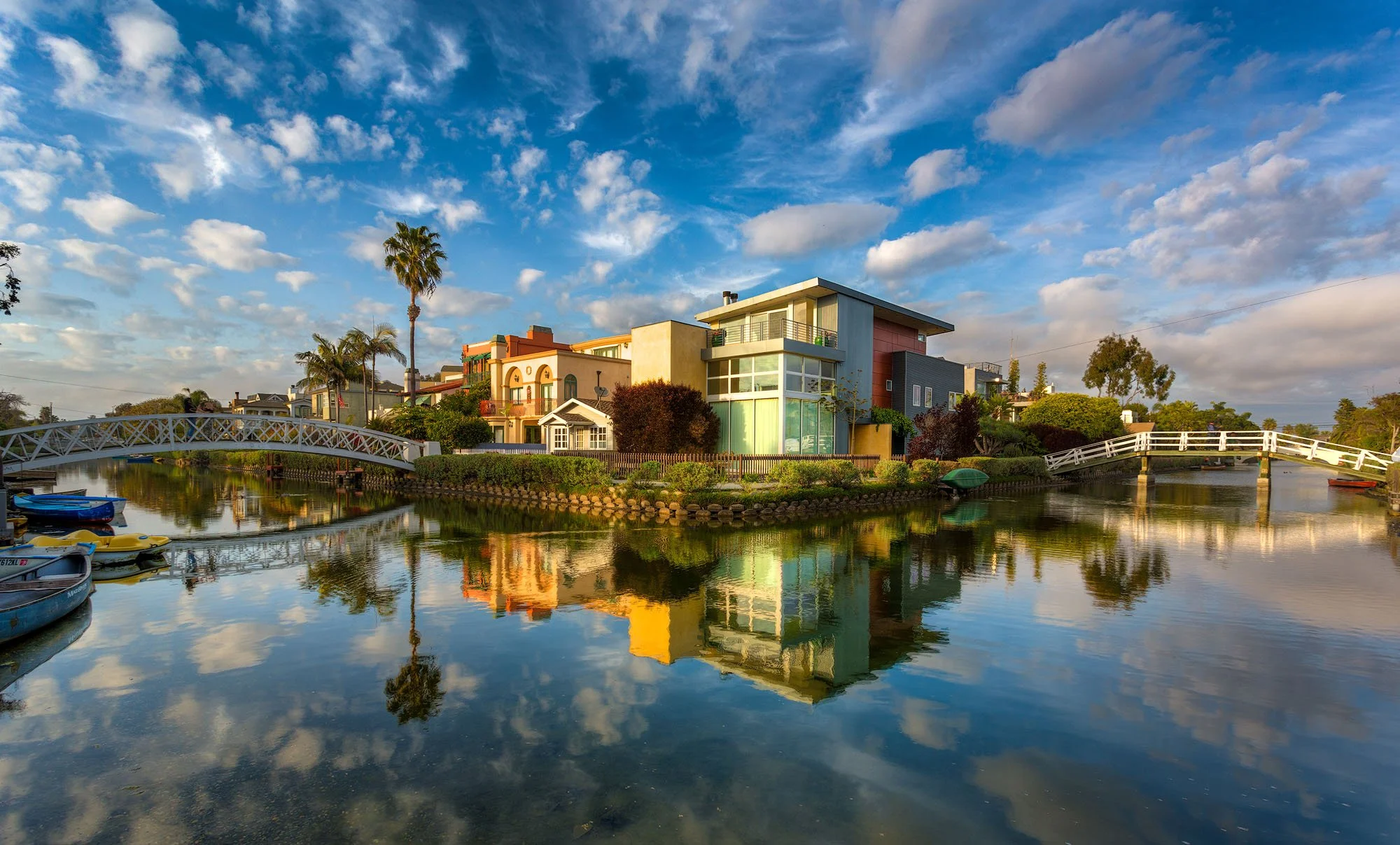 Residential houses along a waterway with small boats, reflected in the calm water, under a partly cloudy sky.