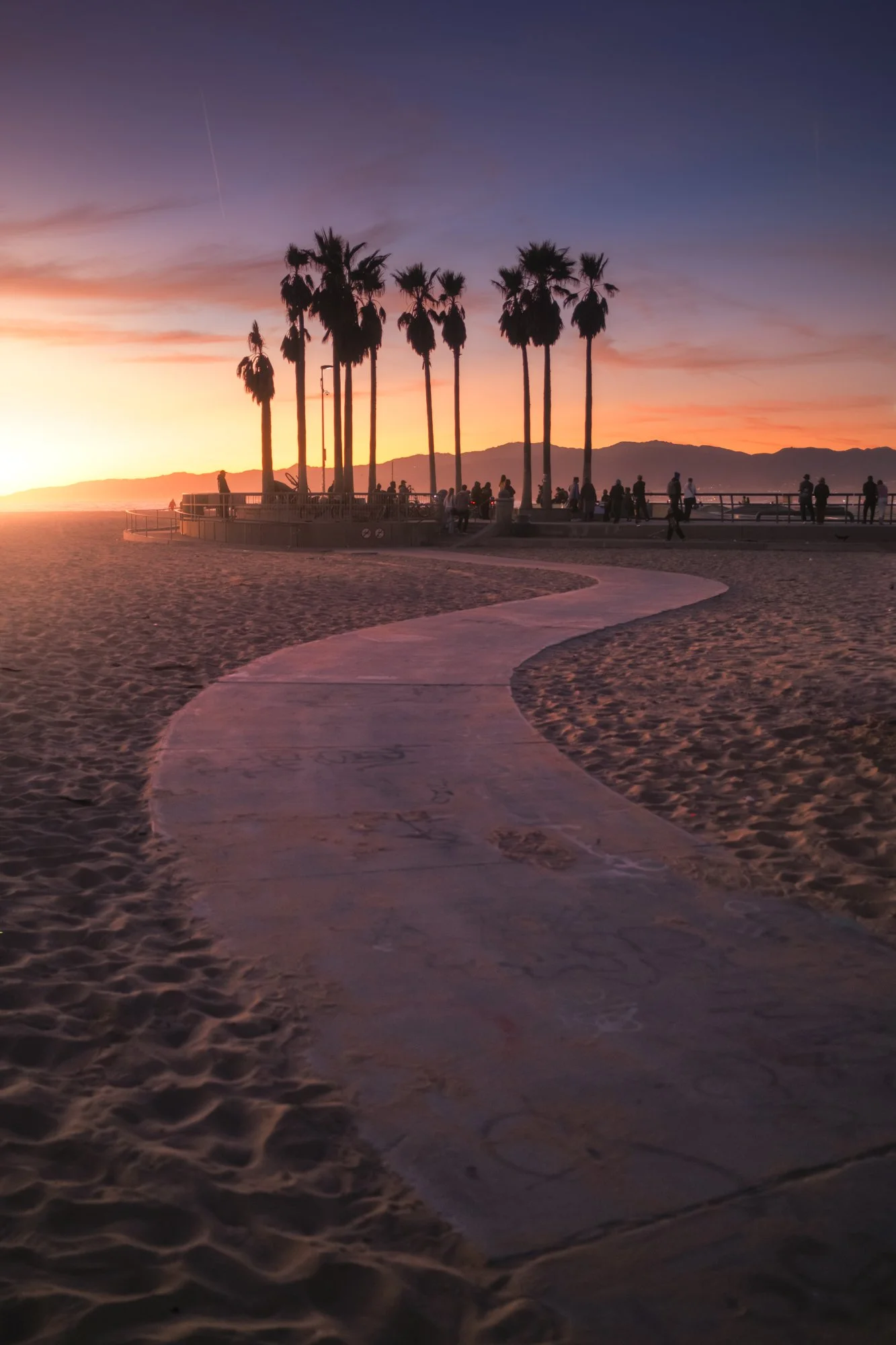Sunset at a beach with a winding pathway leading toward a group of tall palm trees and people near a railing, with mountains in the background.