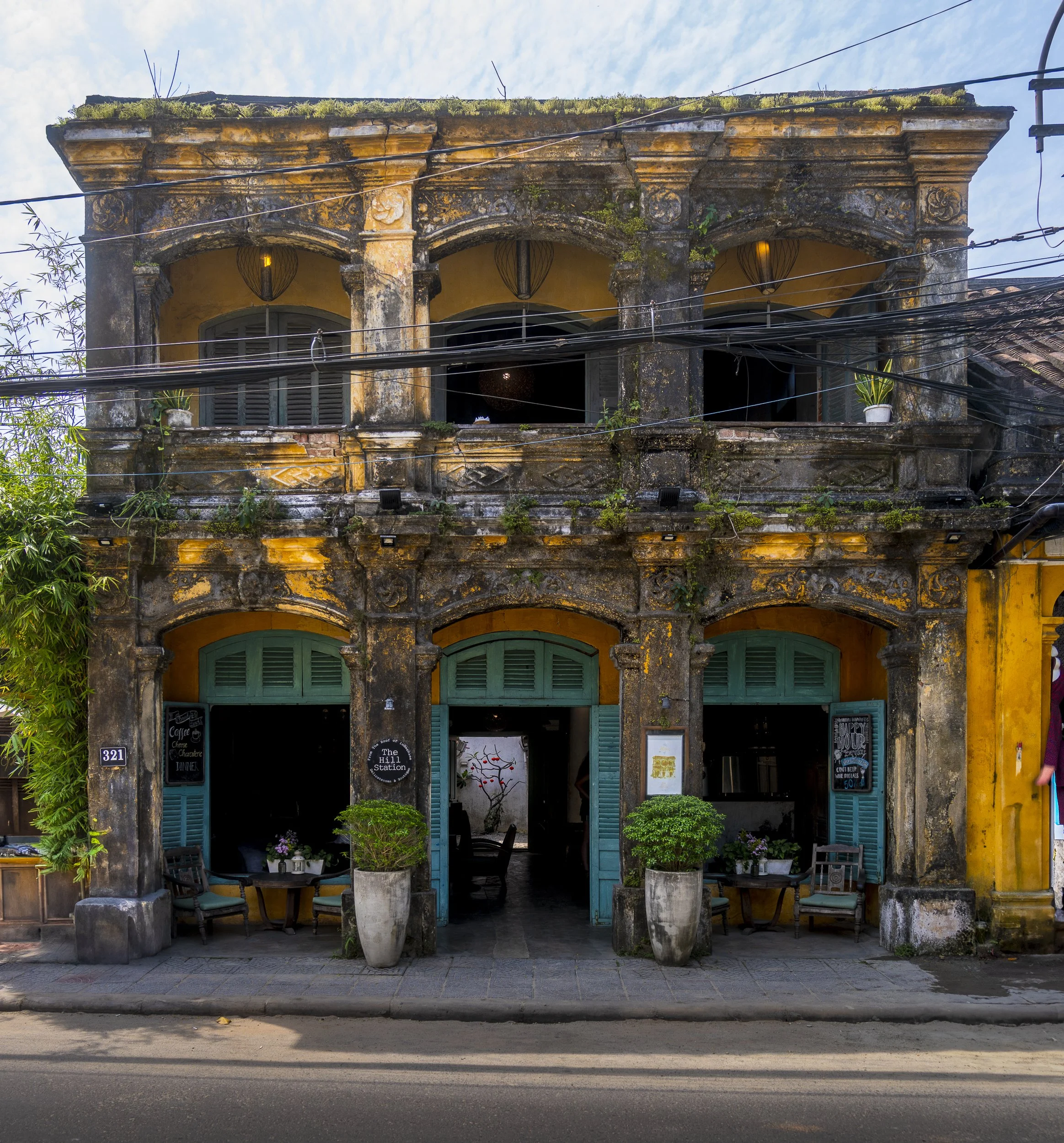 Old two-story building with weathered yellow and dark exterior, green shutters, and a patio with potted plants and outdoor seating.