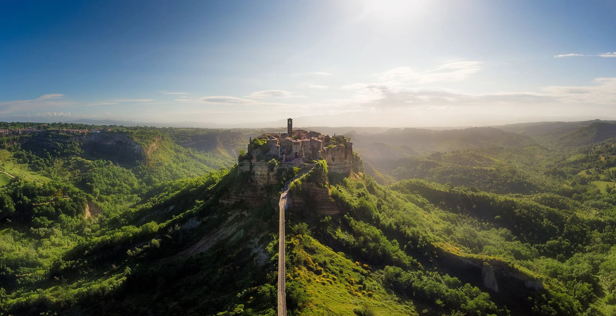 A hilltop town with stone buildings and a clock tower connected by a hanging bridge, surrounded by lush green valleys and hills under a partly cloudy sky.