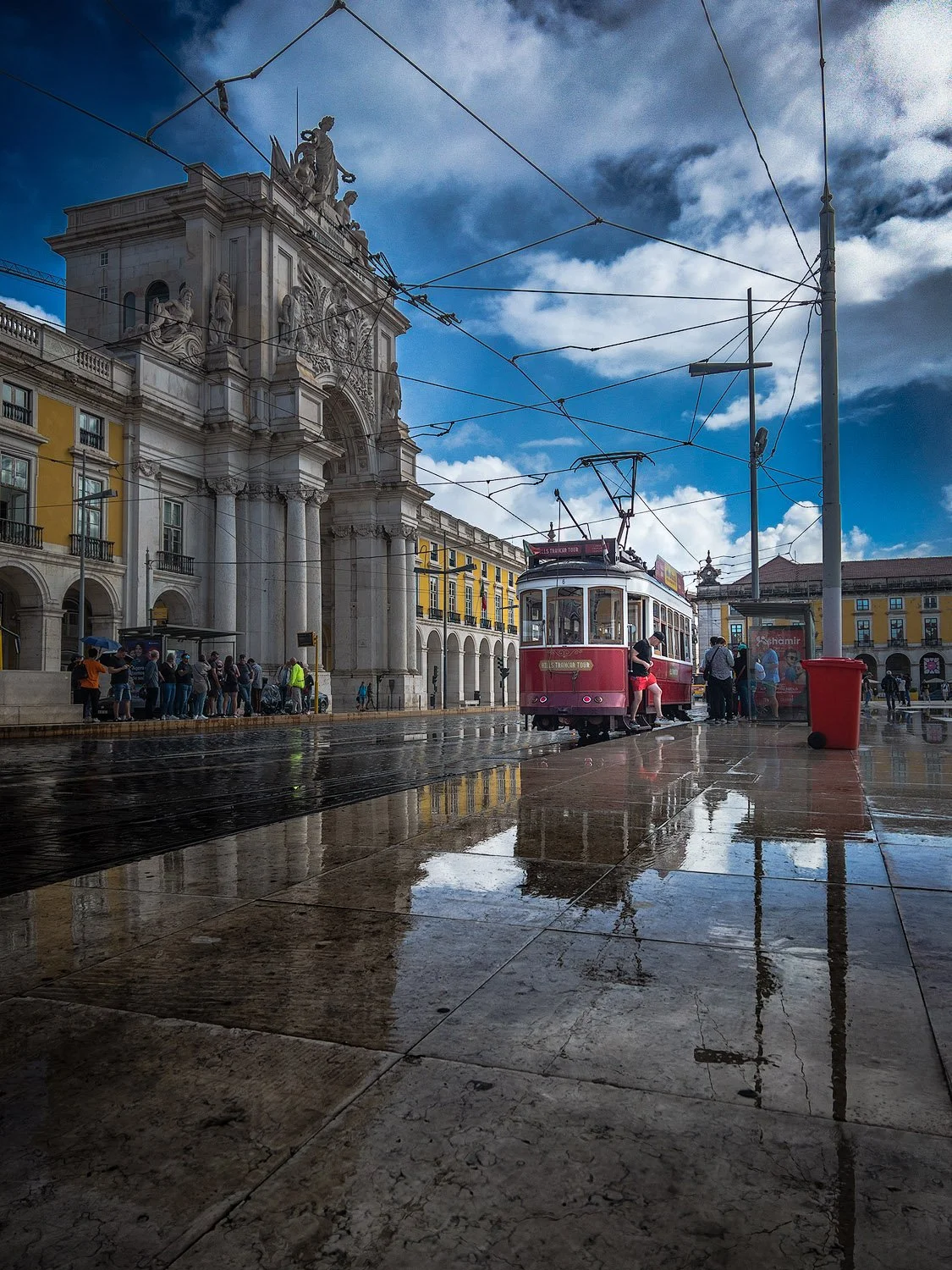 A vintage pink tram at a city square, with a grand historic building featuring statues and ornate architecture in the background. The ground is wet, reflecting the tram and surrounding structures, and a person is stepping off the tram.
