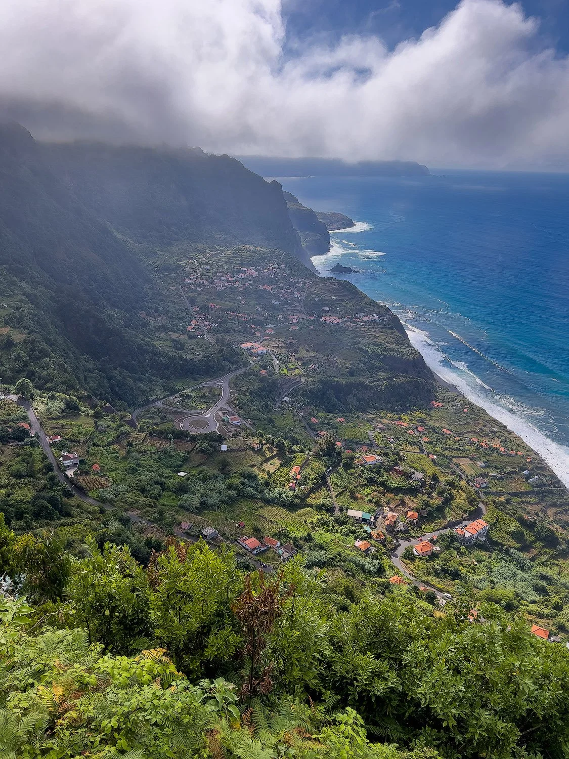 Aerial view of a coastal landscape with green vegetation, winding roads, small houses, a rugged mountain range, and the ocean under a partly cloudy sky.