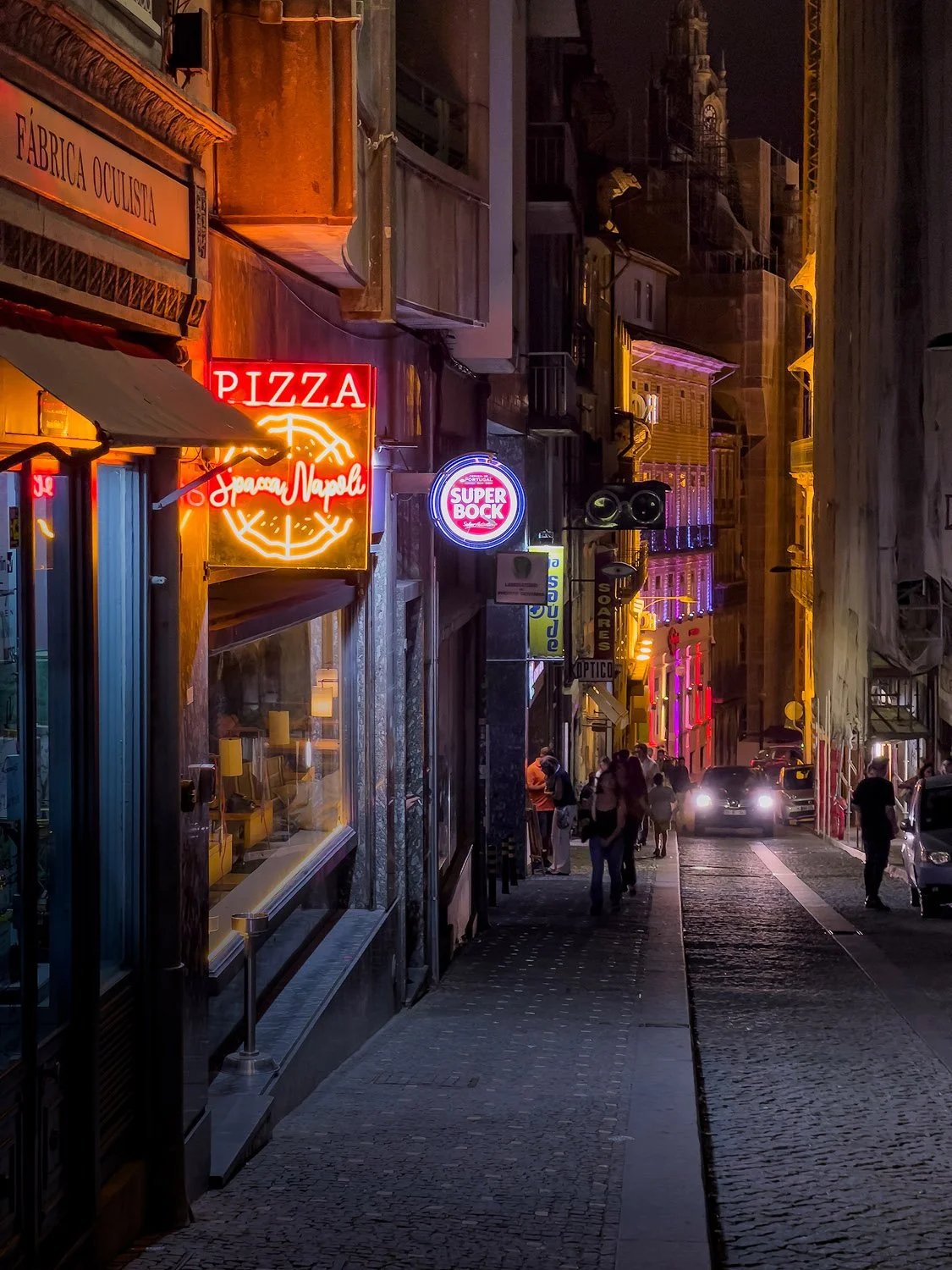 A cobblestone street at night with colorful neon signs for pizza, Super Bock beer, and other businesses, with pedestrians and cars visible in the distance.