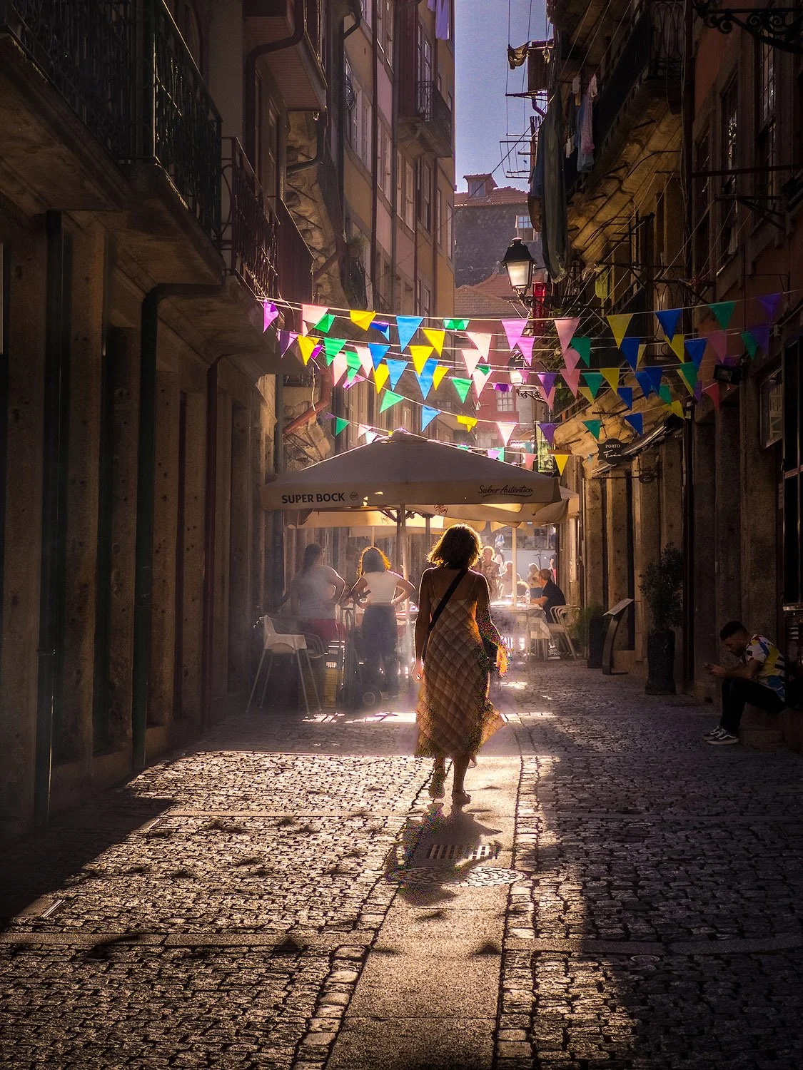 A woman walking down a cobblestone street decorated with colorful triangular flags hanging above. The street is lined with buildings, some with balconies and laundry hanging. There are people sitting and standing at outdoor tables and a person on a b