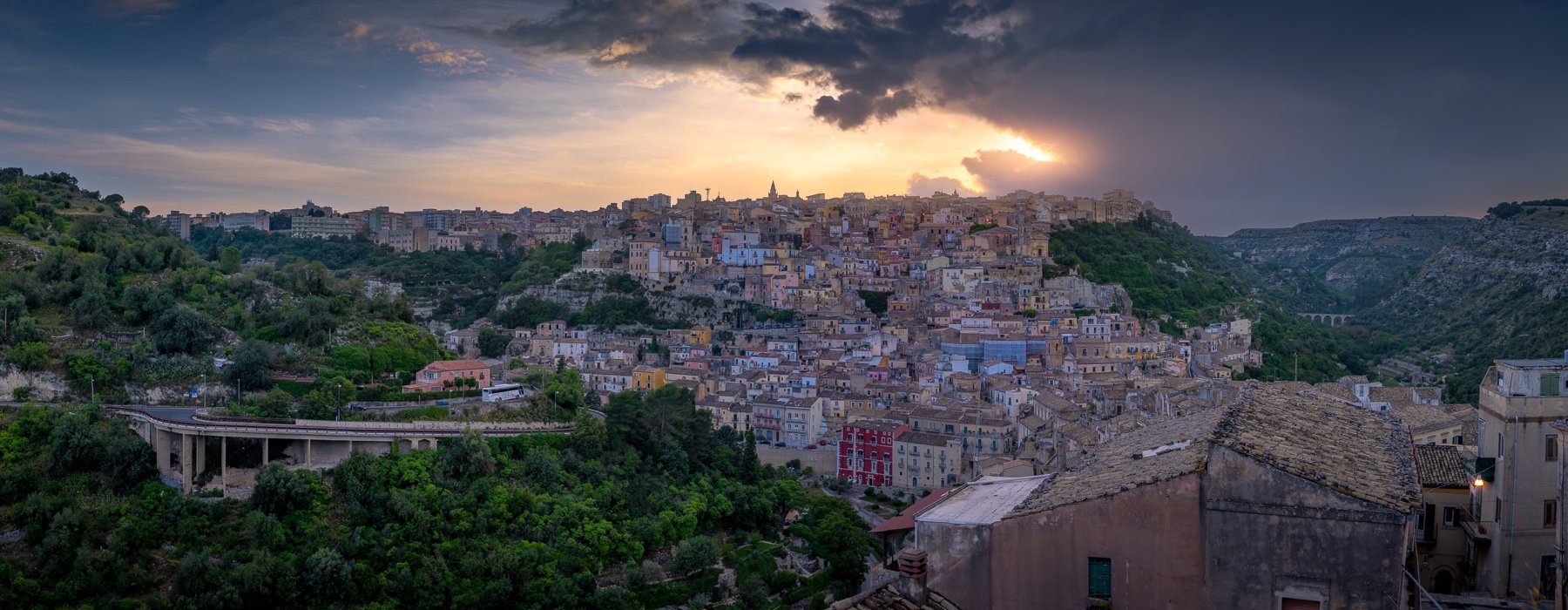 Hilly cityscape at sunset with colorful buildings and green vegetation on the hills.