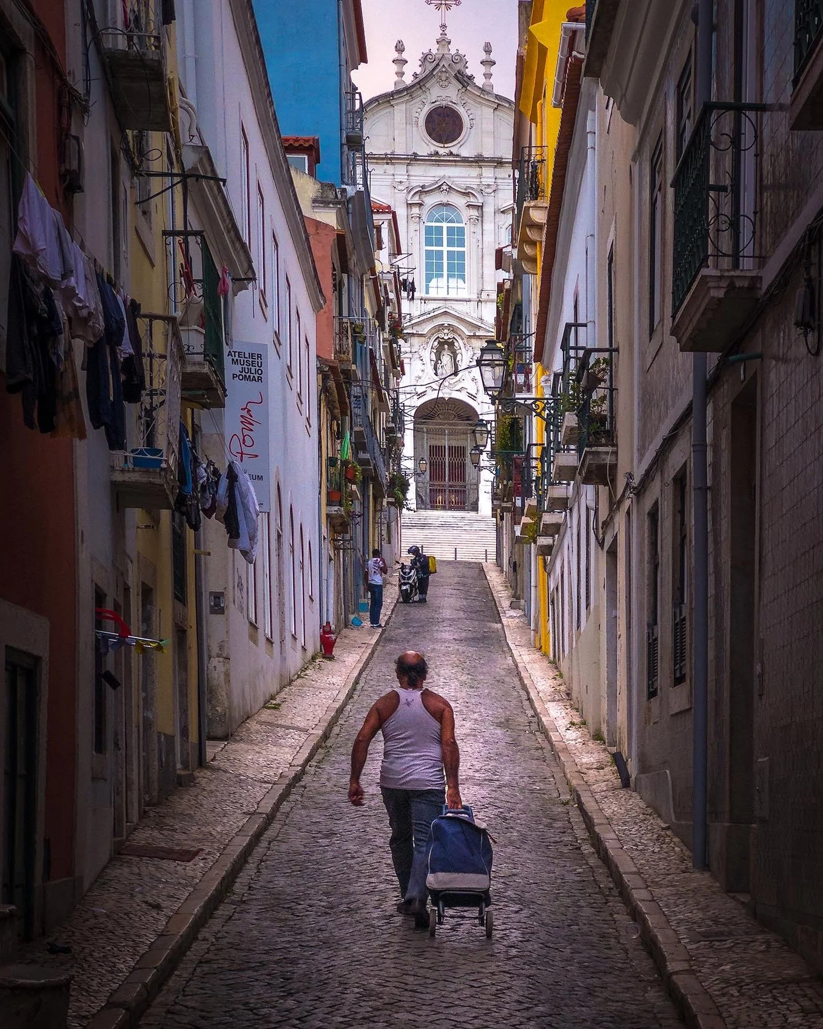 A man walking up a narrow, cobblestone street with a shopping cart in a historic European neighborhood, with colorful buildings and laundry hanging outside on the left. A church or historic building with ornate architecture is visible at the top of t