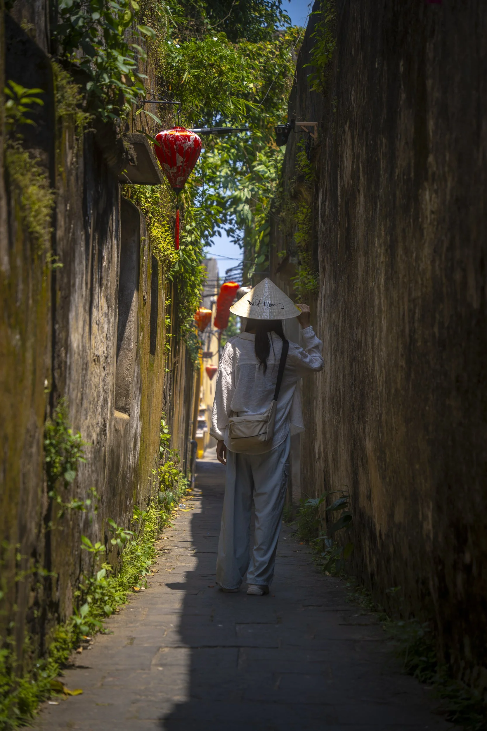 A person walking down a narrow alleyway with traditional red lanterns hanging on the left side, wearing a conical hat and white clothing, with green foliage overhead and sunlight casting shadows.