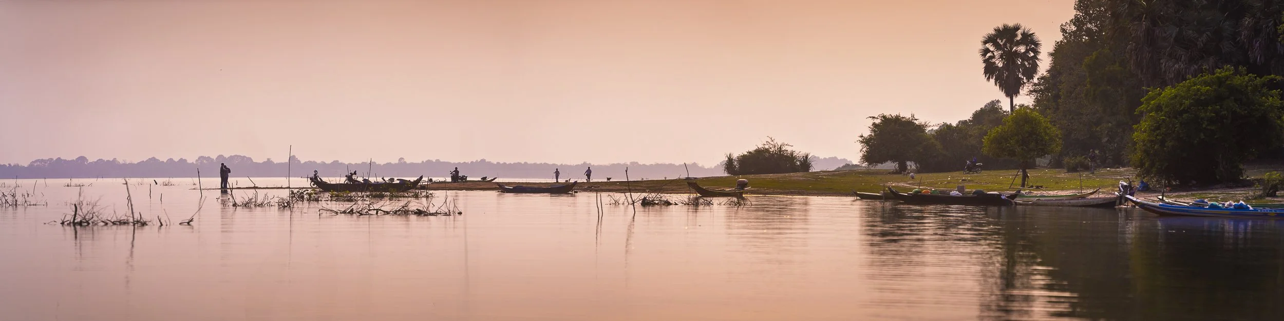 A serene river scene at sunset with boats docked along the bank, trees, and a few people standing and sitting near the water.