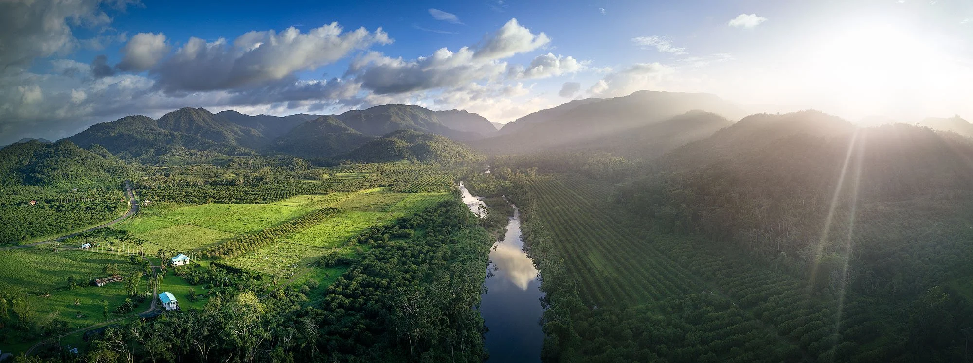 A lush green valley with farmland, a winding river, and mountains in the background under a partly cloudy sky at sunrise or sunset.