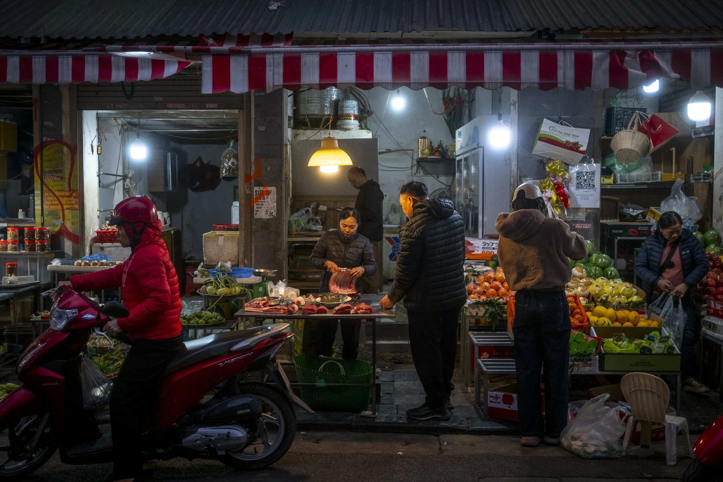 Night scene of a small street market with vendors selling meats and fruits. Several people are shopping, and a woman on a red scooter is passing by. The market is illuminated by hanging lights, with various produce and goods displayed on tables and s