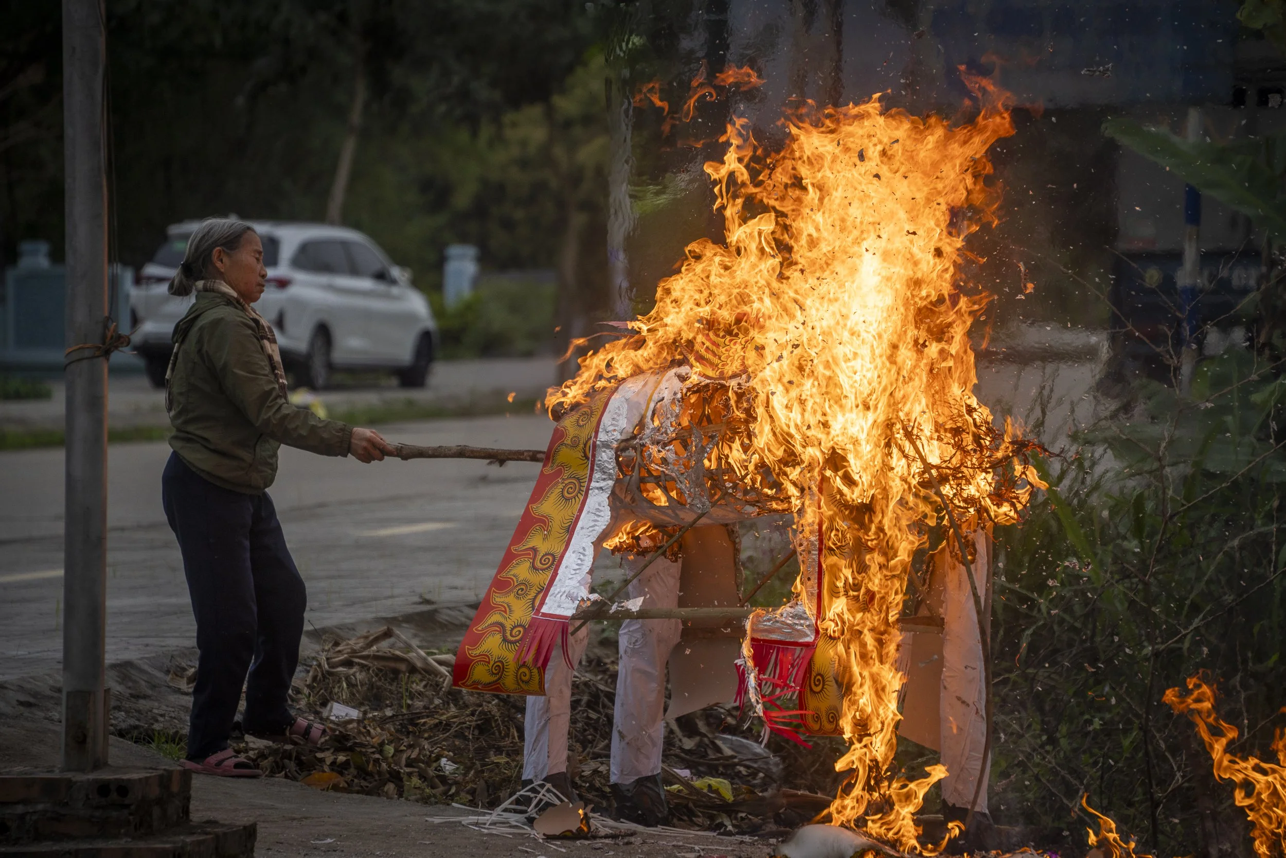 A woman is burning a paper effigy on the side of a street at dusk, with flames engulfing the effigy.