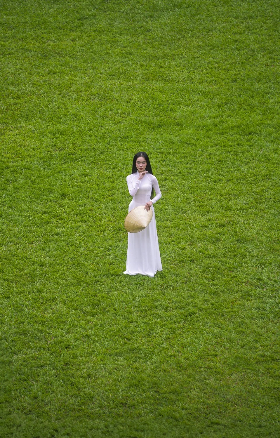 A woman in a long white dress holding a conical straw hat standing on green grass.