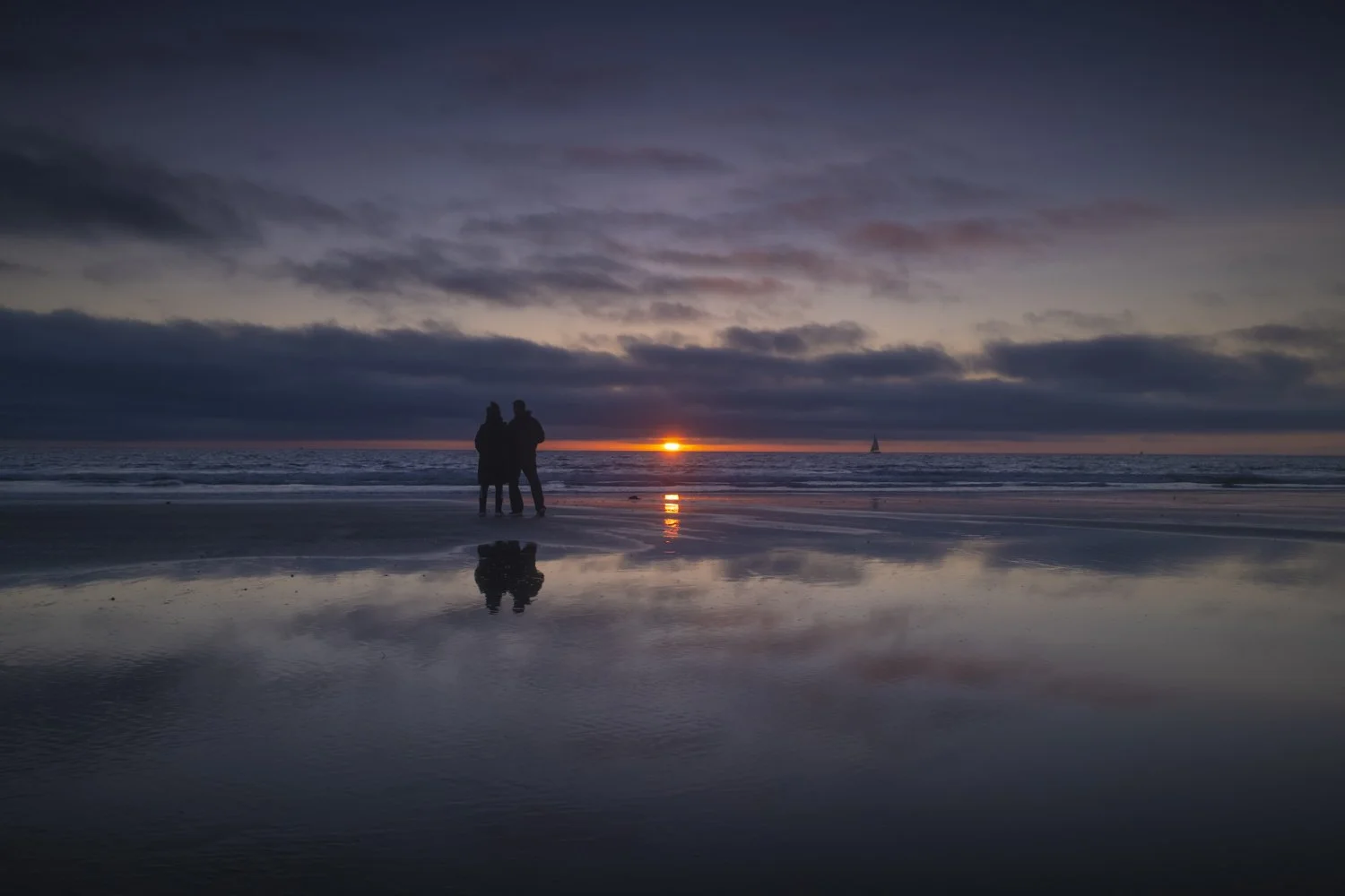 Silhouette of two people standing on a beach during sunset, with a sailboat in the distance and clouds in the sky, reflecting in the wet sand.