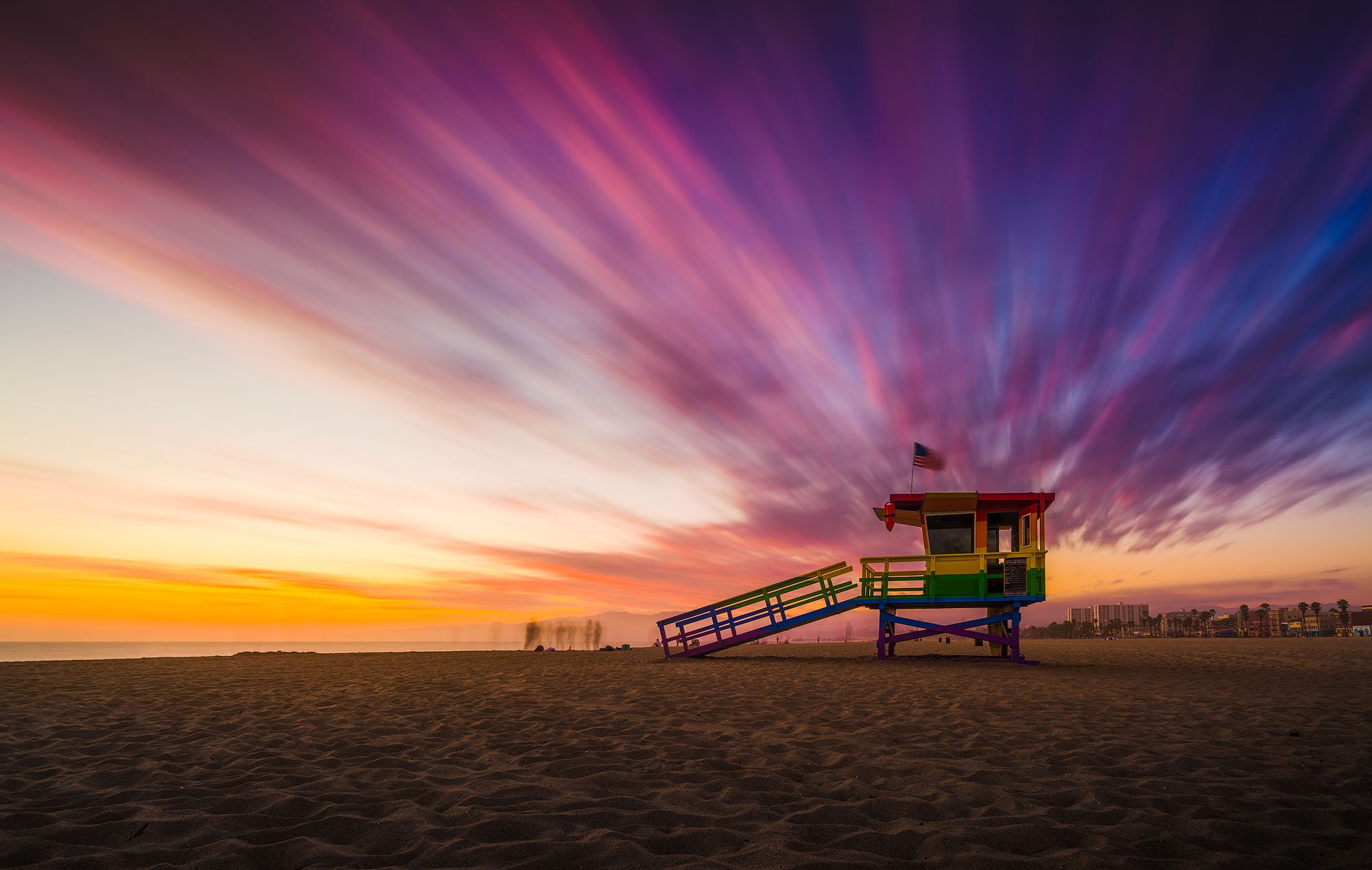 Colorful lifeguard tower on a beach at sunset with vibrant, streaked sky and city buildings in the distance.