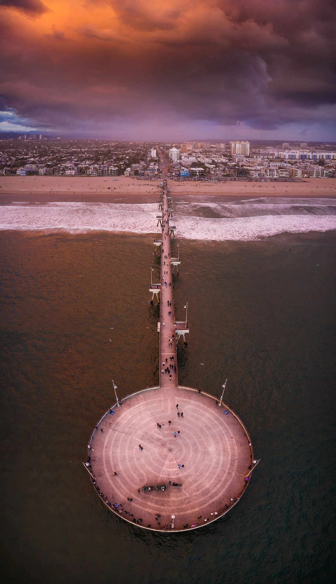 An aerial view of a pier extending into the ocean with a circular platform at the end, under a colorful, cloudy sky, and a city skyline in the distance.