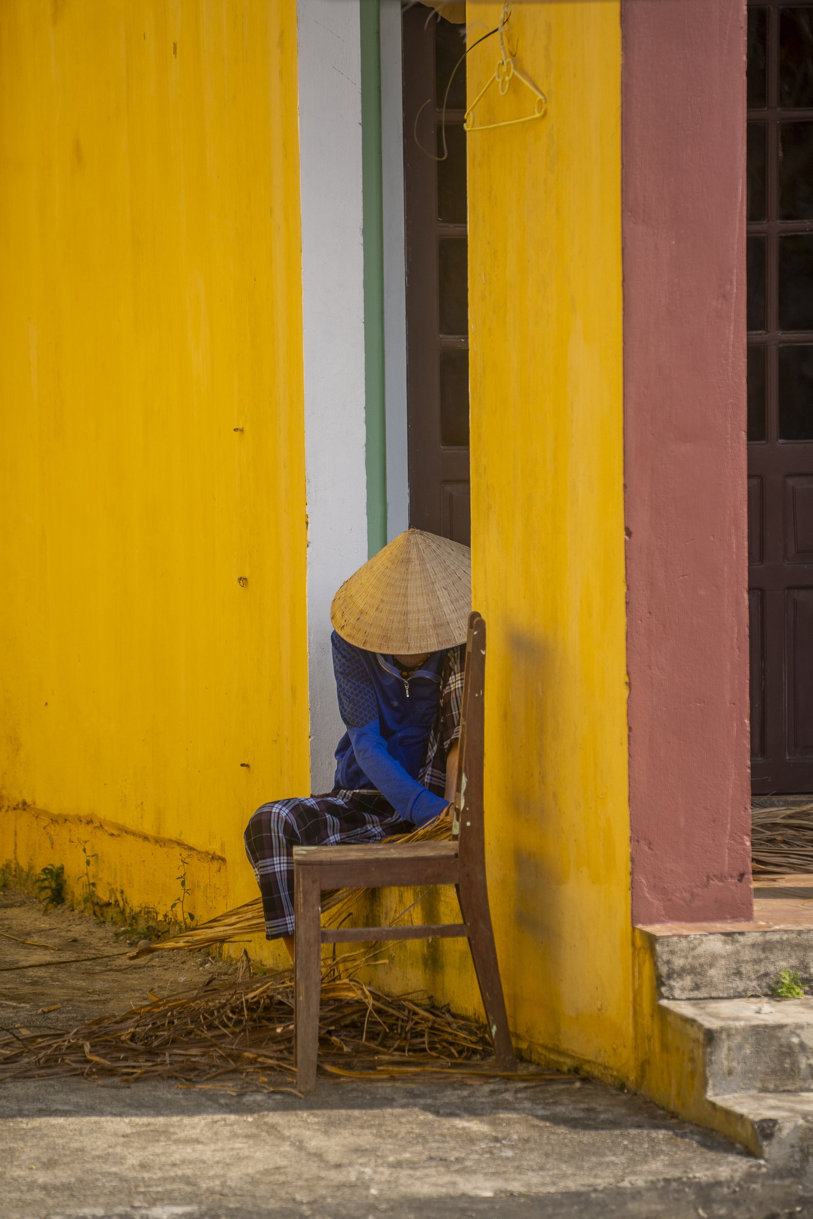 A person sitting on a wooden chair with a traditional conical hat, beside a colorful yellow and pink building with a small staircase.
