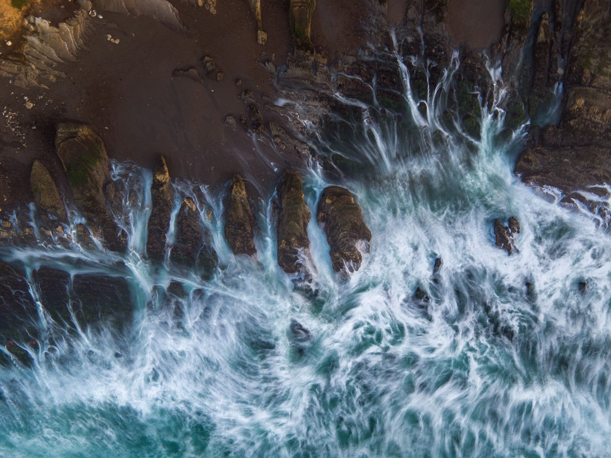 Bird's-eye view of ocean waves crashing against rugged rocky shoreline with spray and foam.