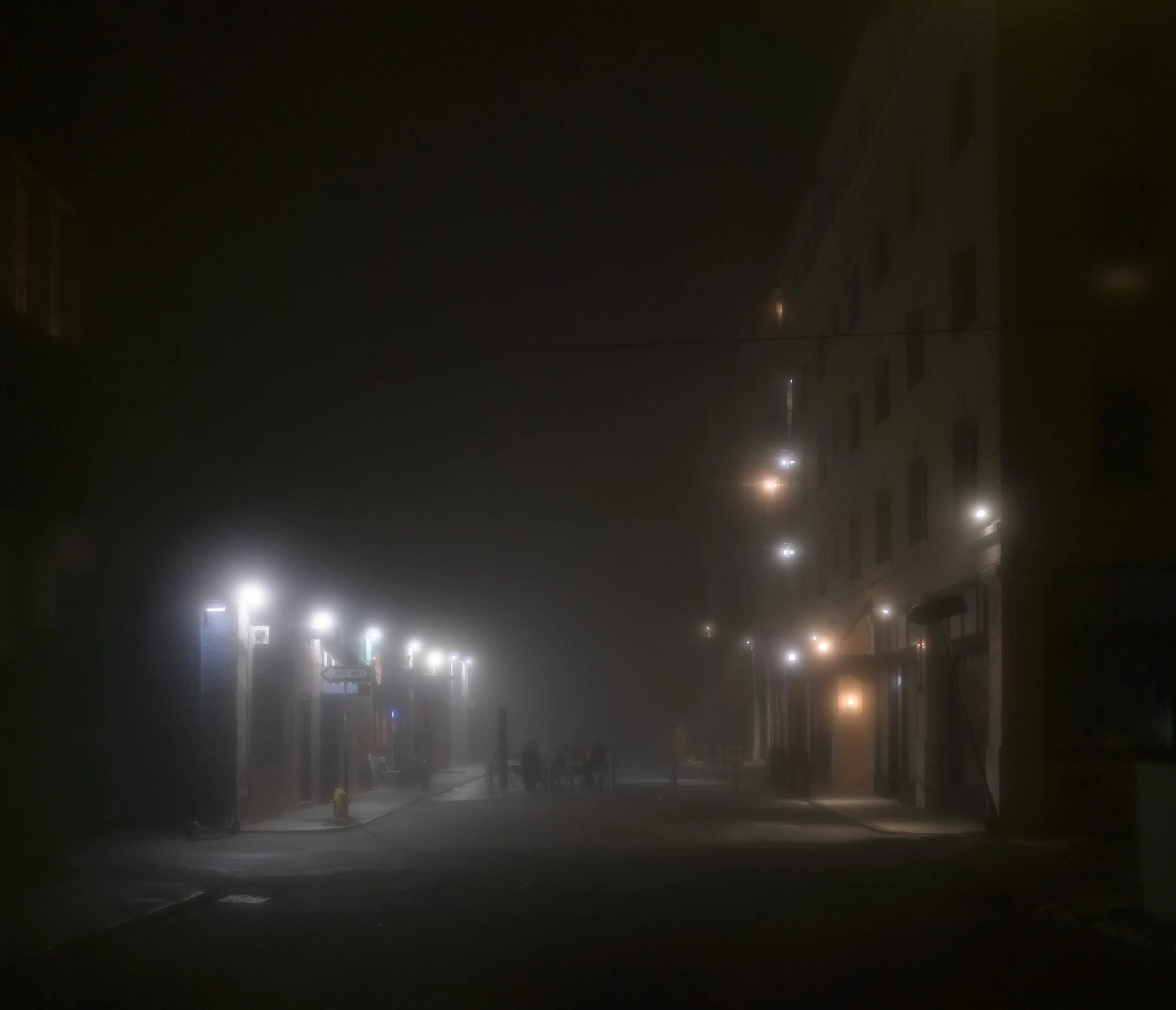 A foggy city street at night with dim streetlights and a few people walking, barely visible through the thick fog.