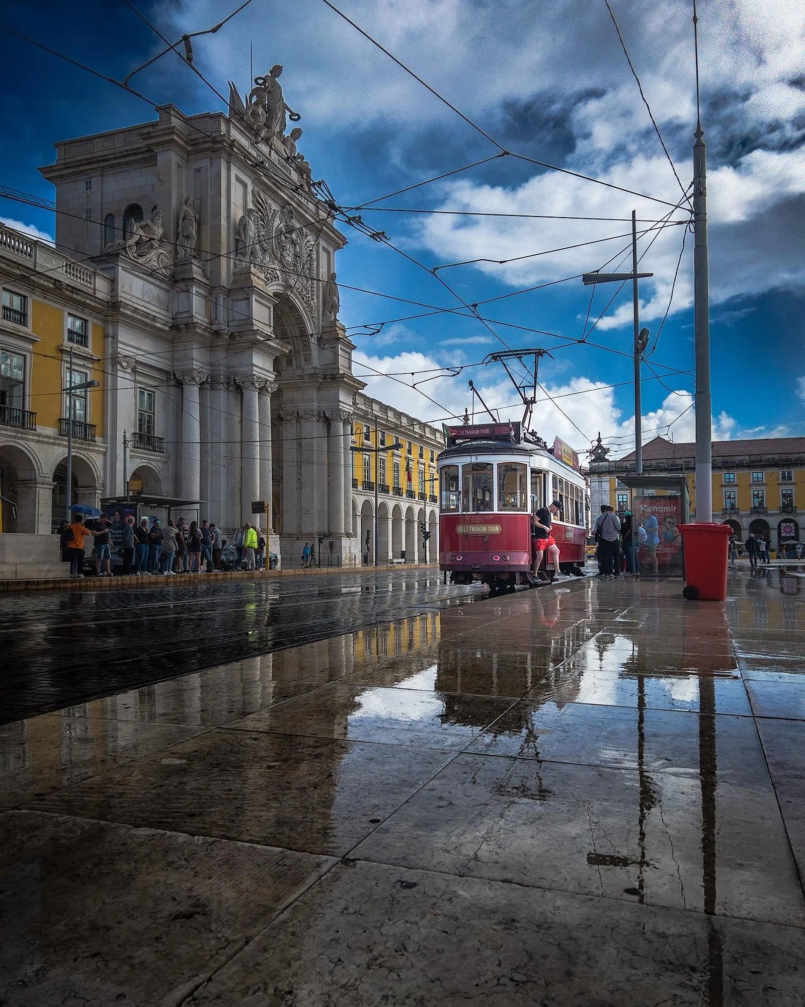 A vintage tram on a rainy day with reflections on wet pavement, people waiting at the tram stop, historic architecture, and a cloudy sky.