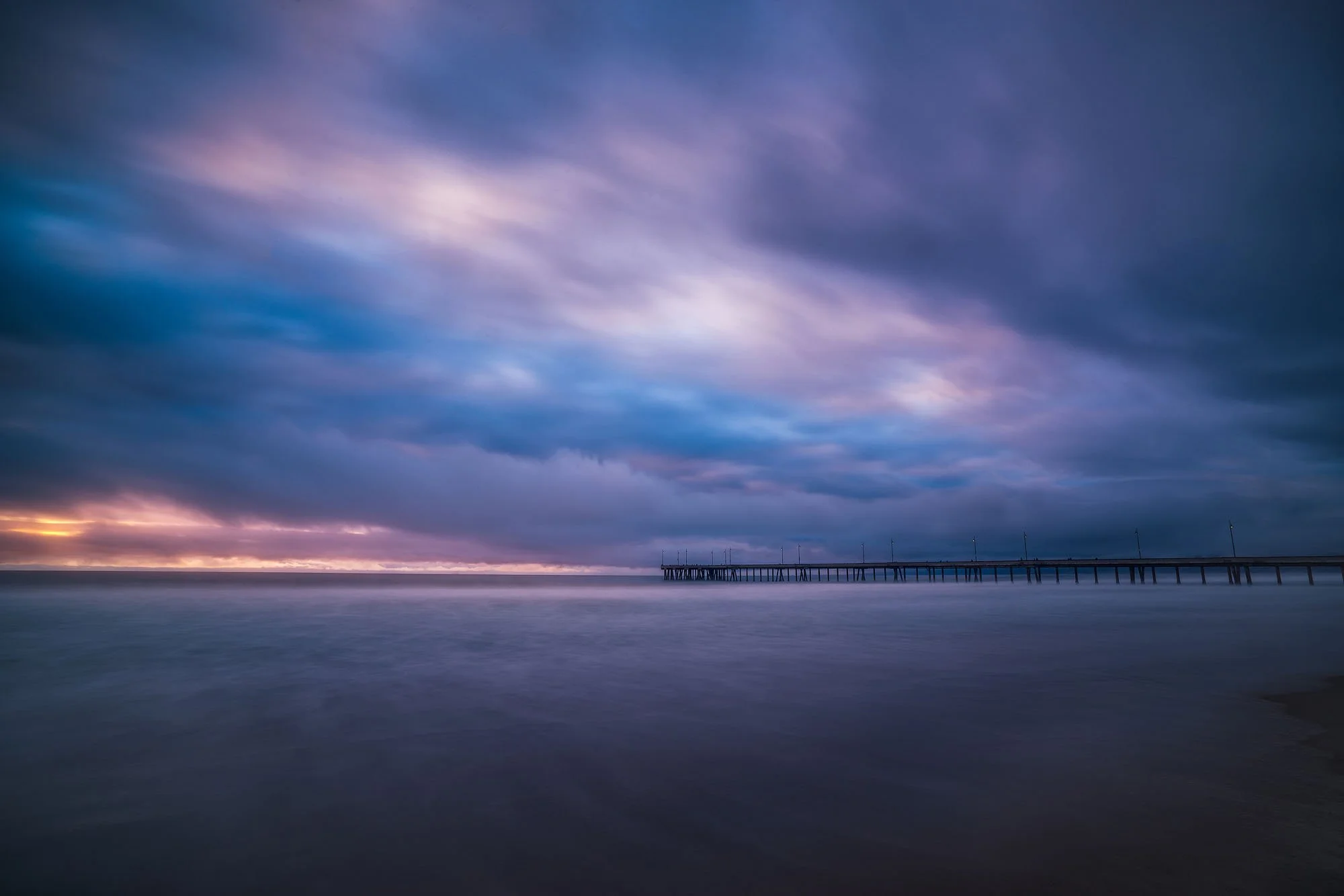 Long exposure photograph of a pier extending into the ocean during a cloudy sunset, with purple and pink hues in the sky.