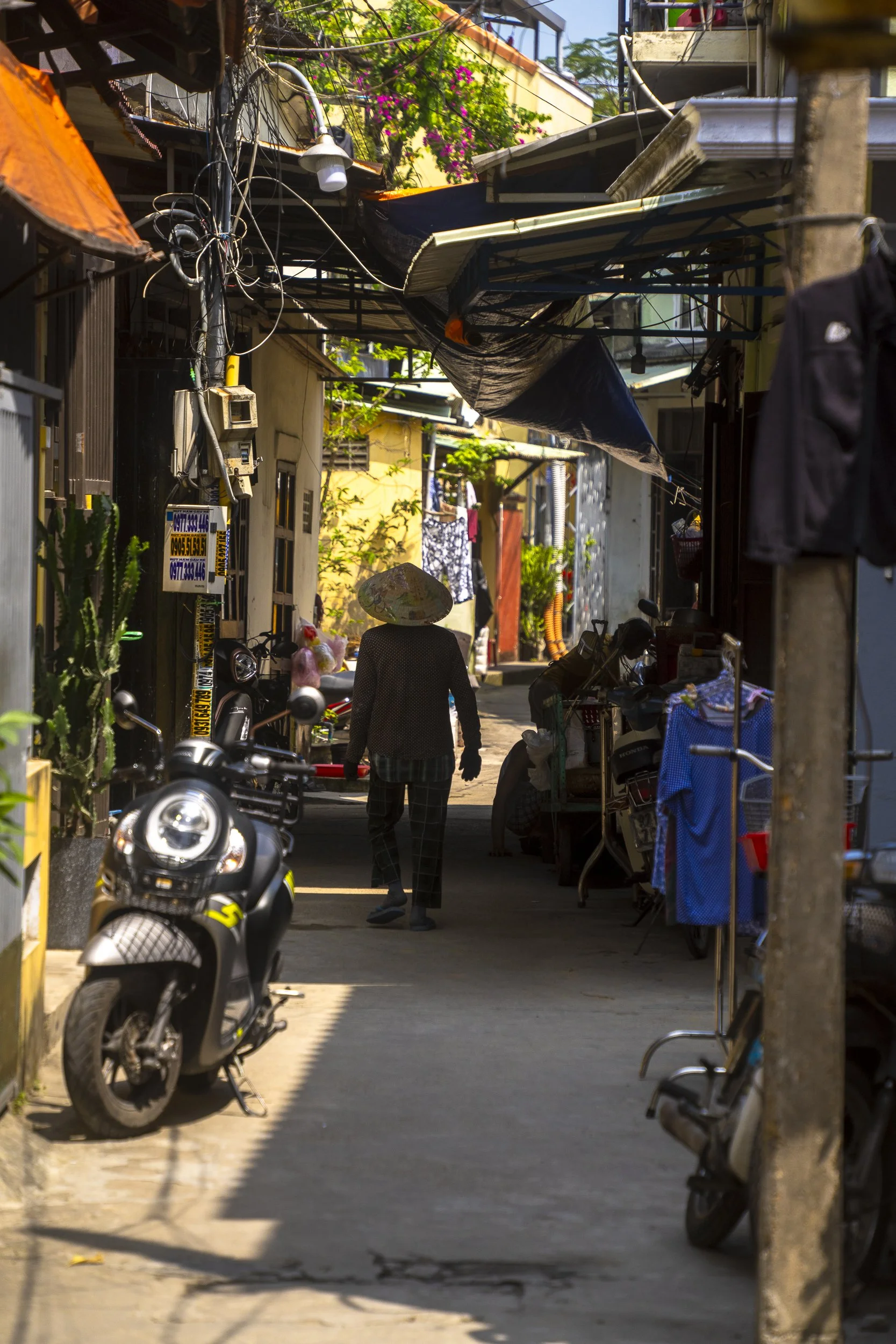 A narrow alleyway in a residential area with motorcycles parked along the sides and clothes hanging to dry. A person wearing a traditional conical hat walks away from the camera. The alley is shaded by makeshift awnings and contains various plants.