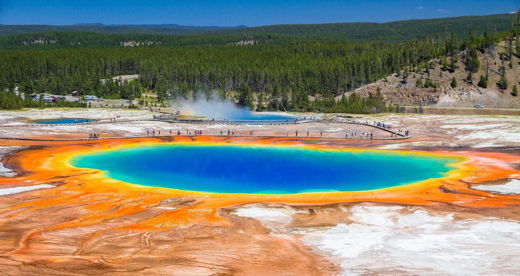 Colorful hot spring with orange, yellow, green, and blue water, surrounded by greenery and a blue sky.