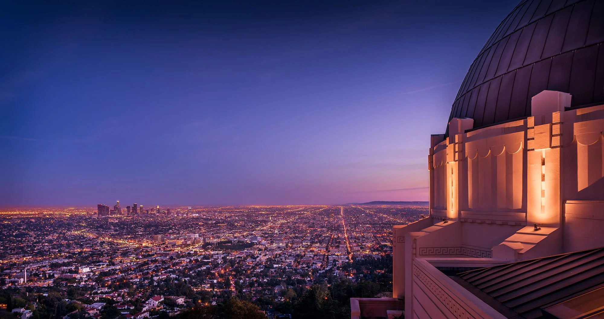 Nighttime cityscape view of Los Angeles from Griffith Observatory with city lights and skyline in the distance, and the observatory's dome in the foreground.