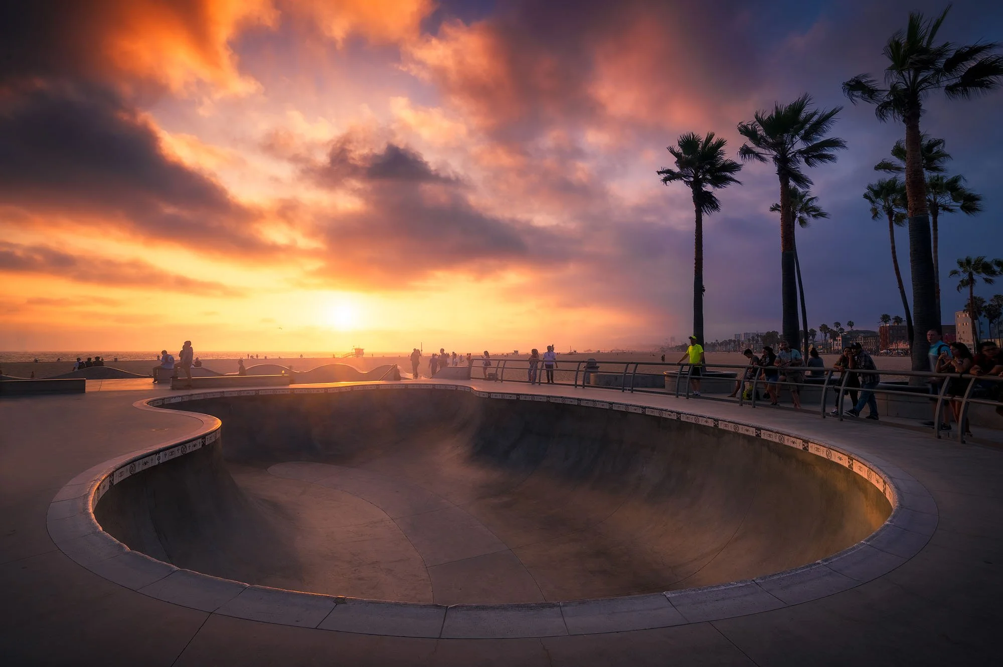A skatepark at sunset with a bowl in the foreground and people walking and sitting along the perimeter. Palm trees line the background with a partly cloudy sky painted in orange and purple hues.