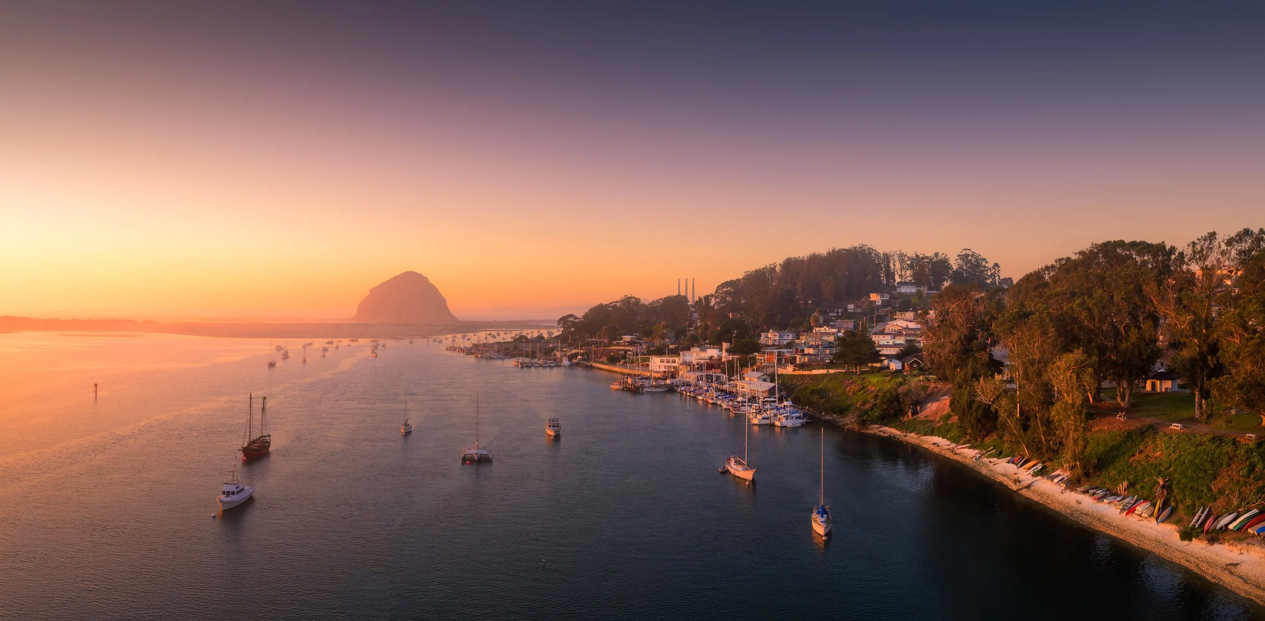 Sunset over a coastal waterside town with boats docked in the harbor, hillside houses, and a distant rock formation.