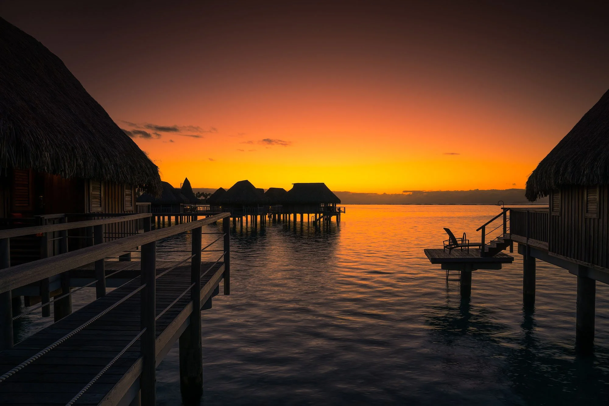 Overwater bungalows with thatched roofs against a vibrant sunset sky, reflecting on calm water.