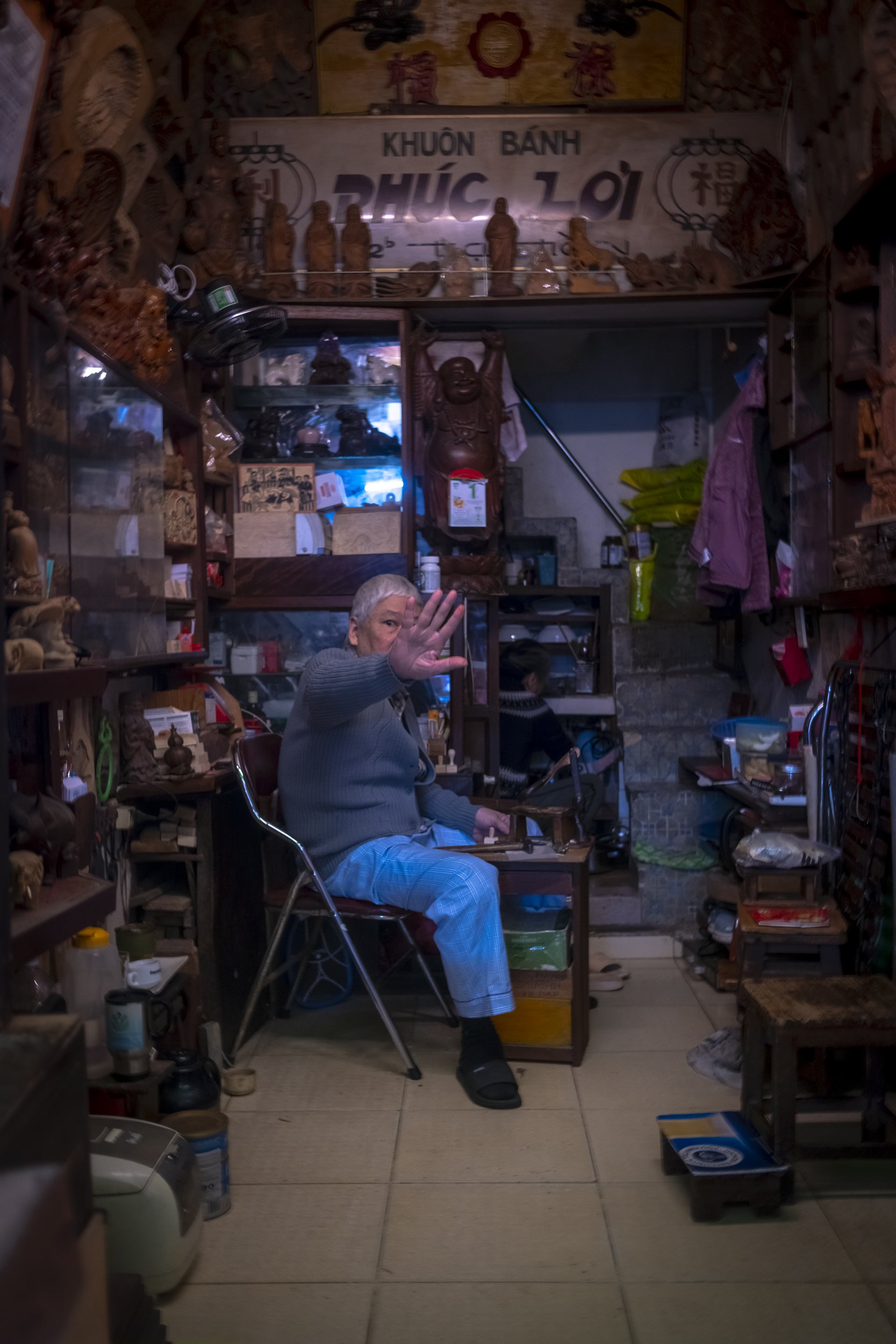 An elderly person with gray hair sitting in a small, cluttered shop, gesturing with an open palm towards the camera. The shop is filled with wooden carvings, books, and various items, with a sign in Vietnamese above reading "Khuôn Bánh" and "Phúc Lợi