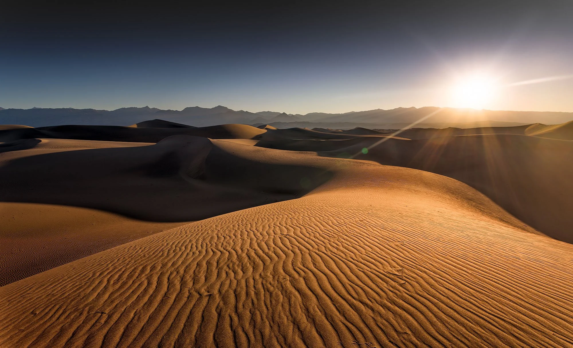Sunset over a desert with rolling sand dunes and distant mountain range.