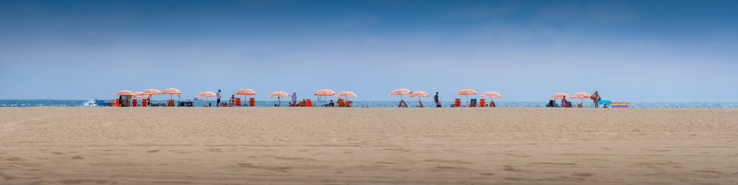 Empty sandy beach with orange-striped umbrellas, beach chairs, and people in the background near the ocean under a clear blue sky.
