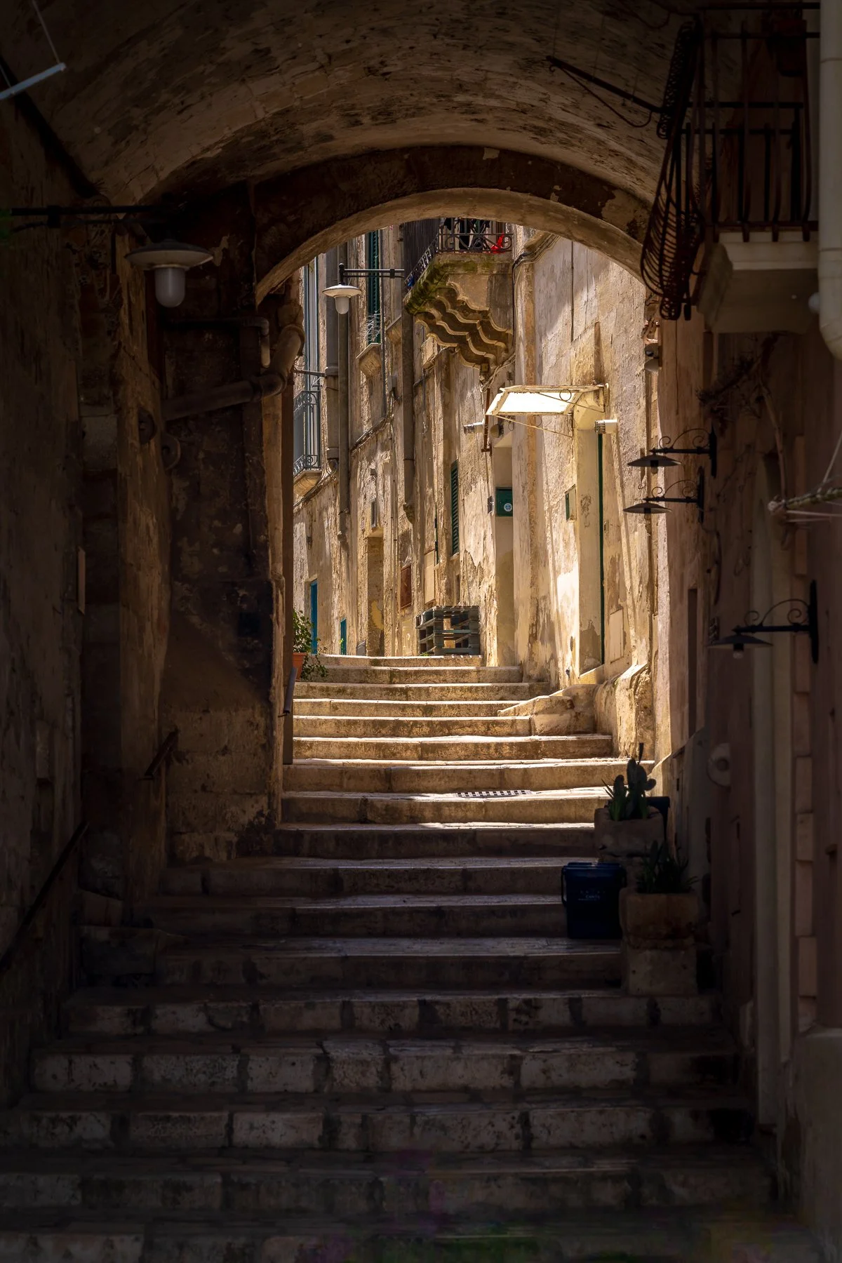 A narrow, sunlit stone staircase in an old European alleyway, flanked by weathered stone walls and small potted plants, with an archway at the top.