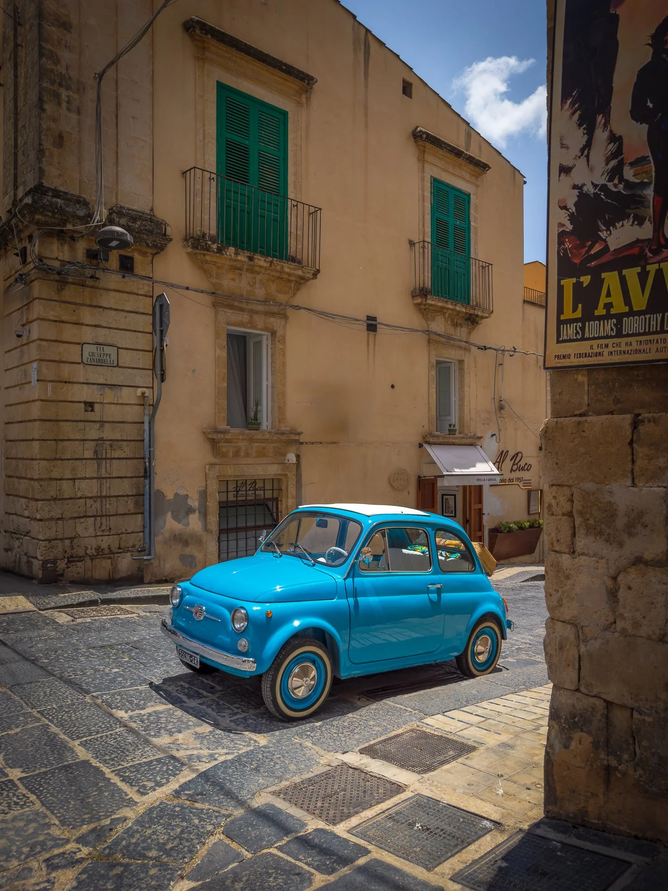 A small blue vintage Fiat 500 car parked on a cobblestone street in a historic European neighborhood with aged yellow buildings, green window shutters, and a partially cloudy sky overhead.