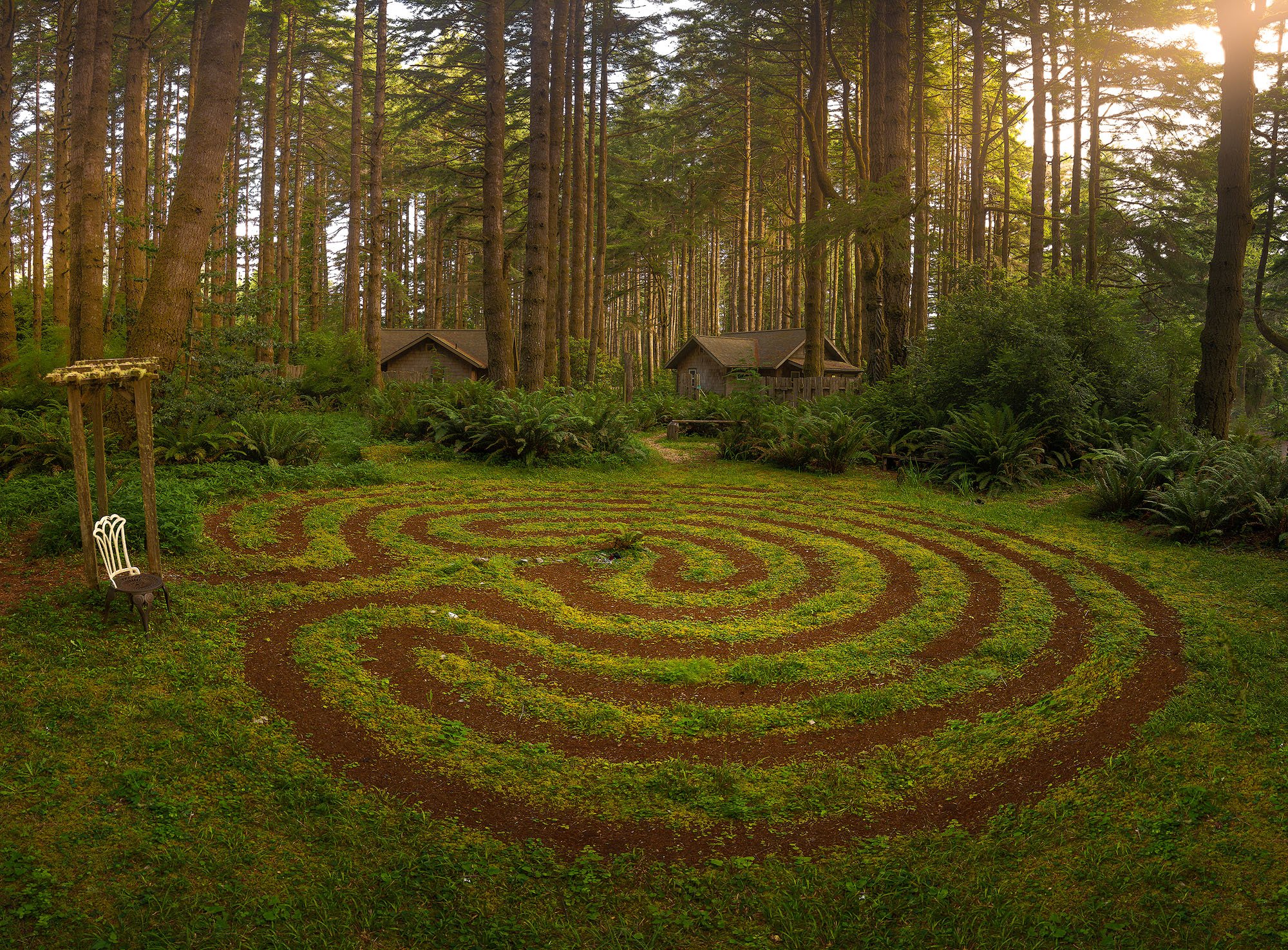 A circular winding garden maze on green grass in a forest clearing, with a swing and a chair on the left, surrounded by tall trees, some with shrubs and small houses in the background, during sunset.
