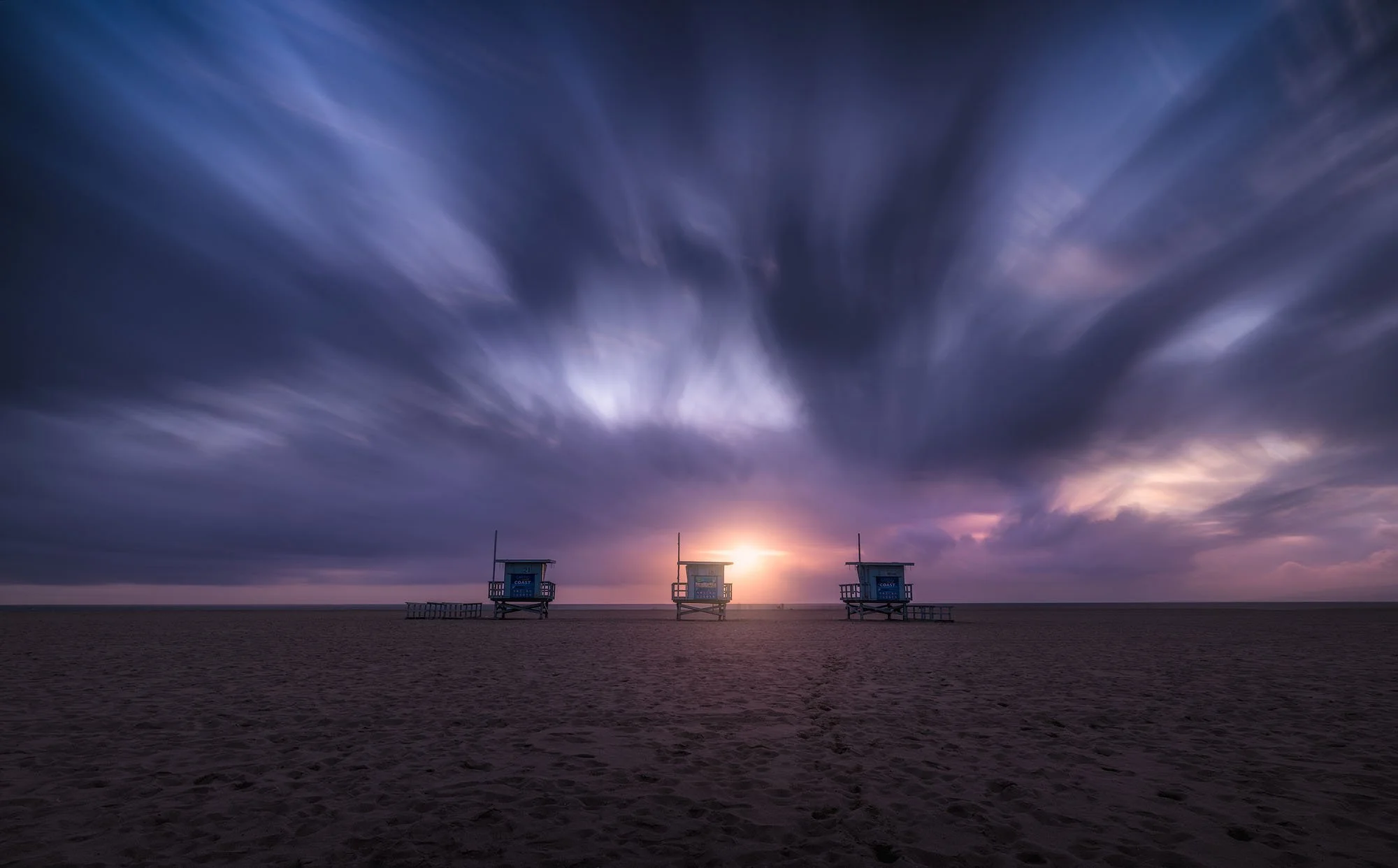 Three lifeguard towers on a beach at sunset with dramatic purple clouds in the sky.