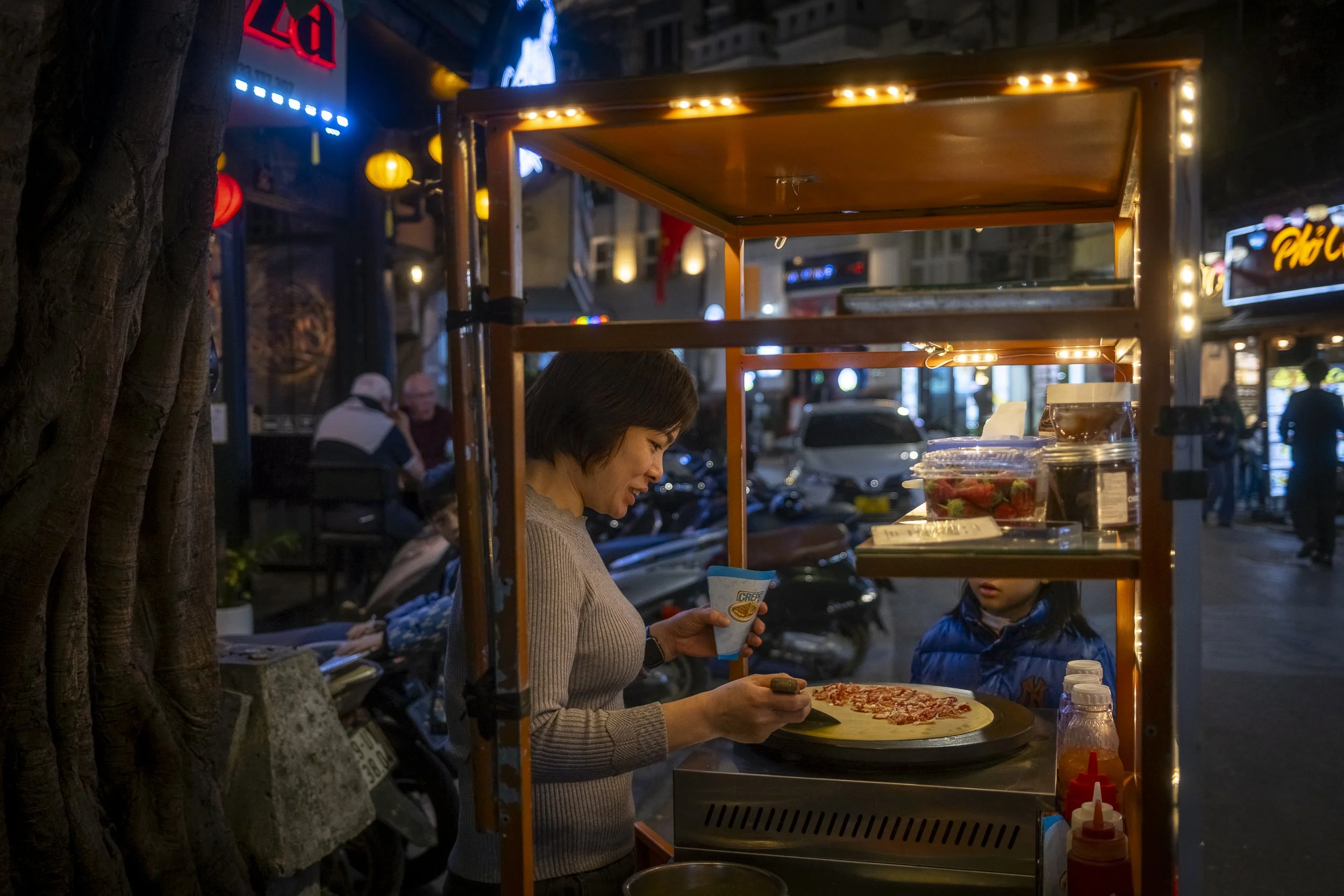 A street vendor prepares a pizza in a cart at night, with city lights and people in the background.
