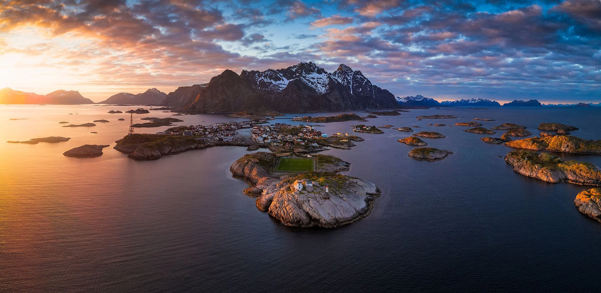 Sunset over a landscape of mountains and small islands with houses, a lighthouse, and a football field on a rocky peninsula in a calm body of water.
