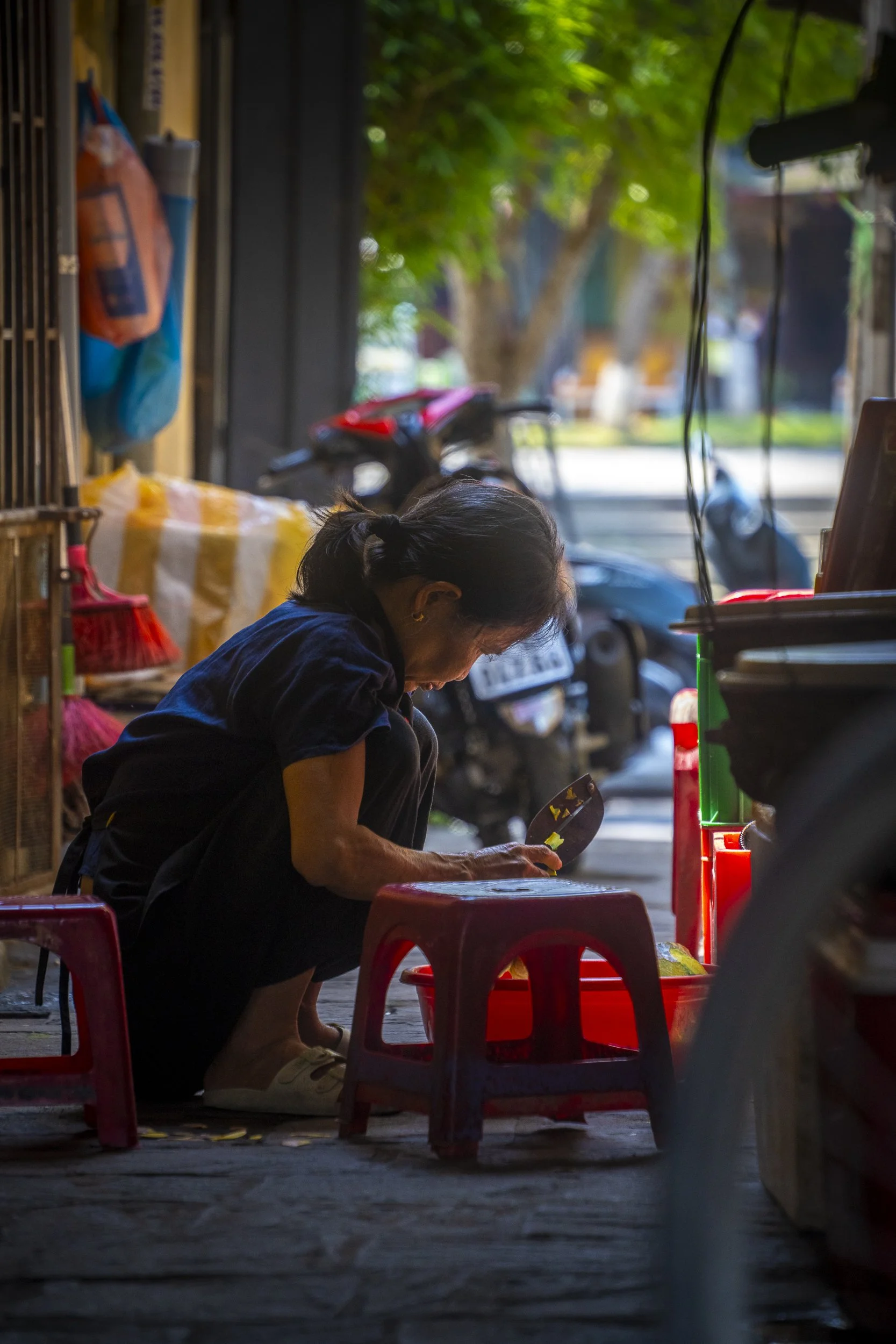 An elderly woman crouching on a small plastic stool, looking at her phone, with a busy street scene in the background.