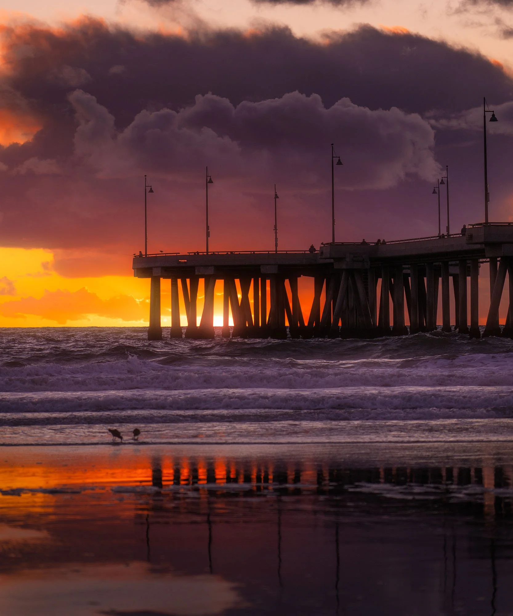 Sunset on a beach with a pier extending into the water, dark clouds overhead, and reflections in the wet sand.