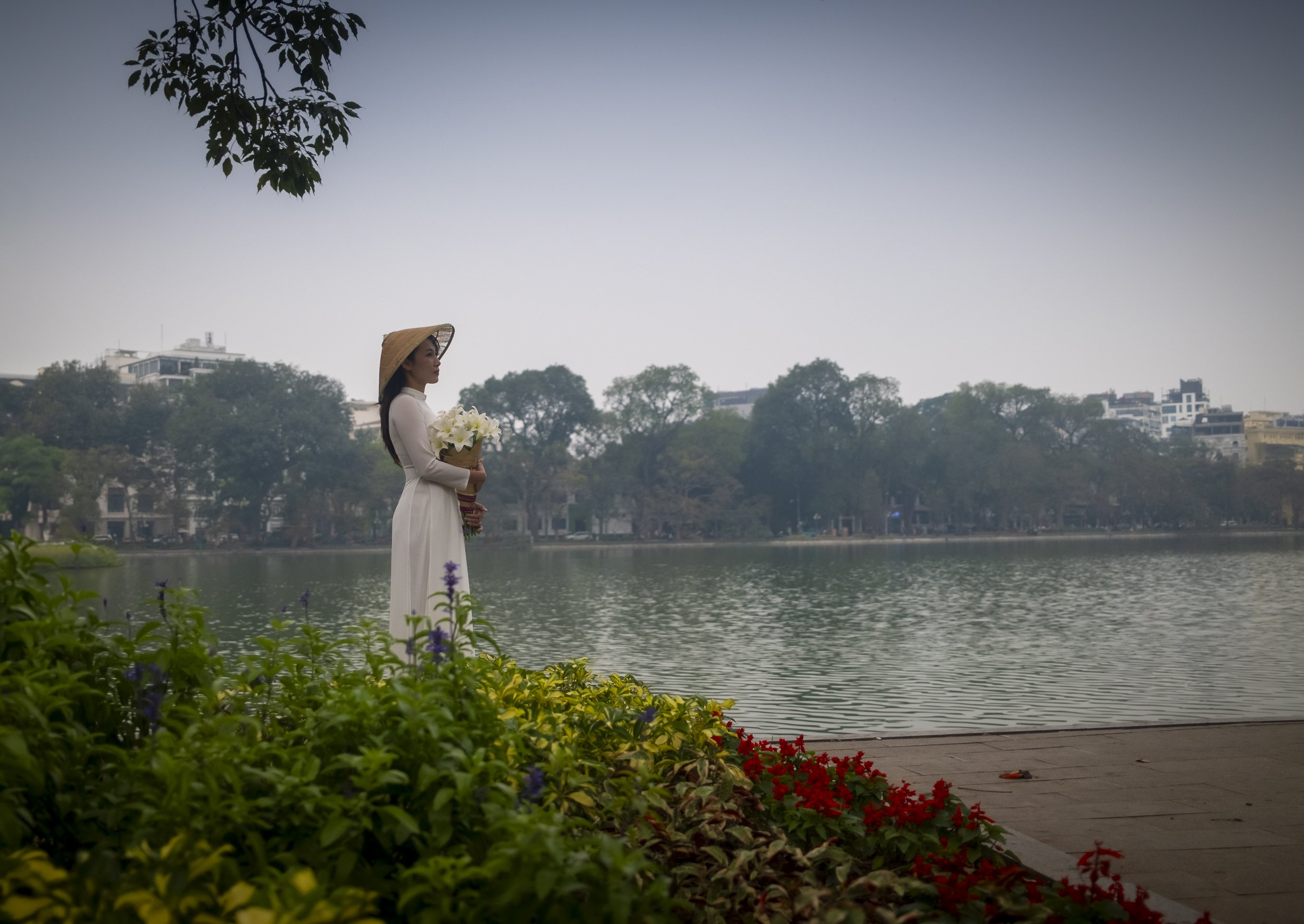 A woman in a white dress and straw hat holding a bouquet of white flowers by a lake, with colorful flowers in the foreground and trees and buildings in the background.