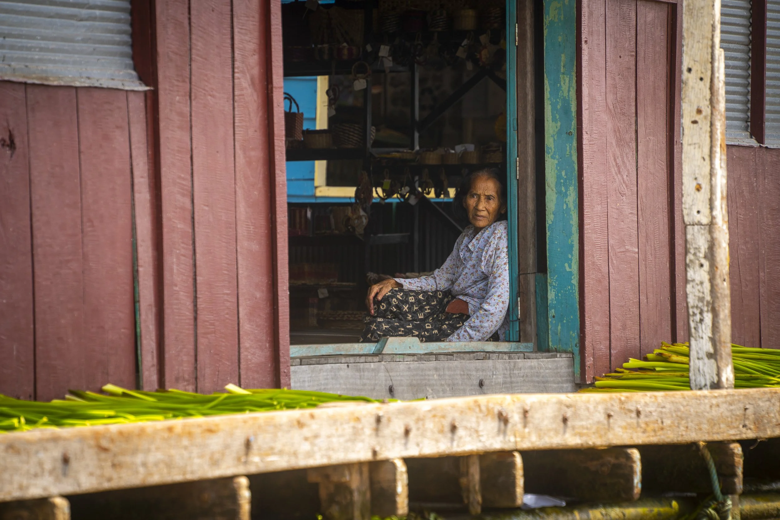An elderly woman sitting inside a wooden house, looking out through an open window, with green bananas laid outside on a wooden platform.