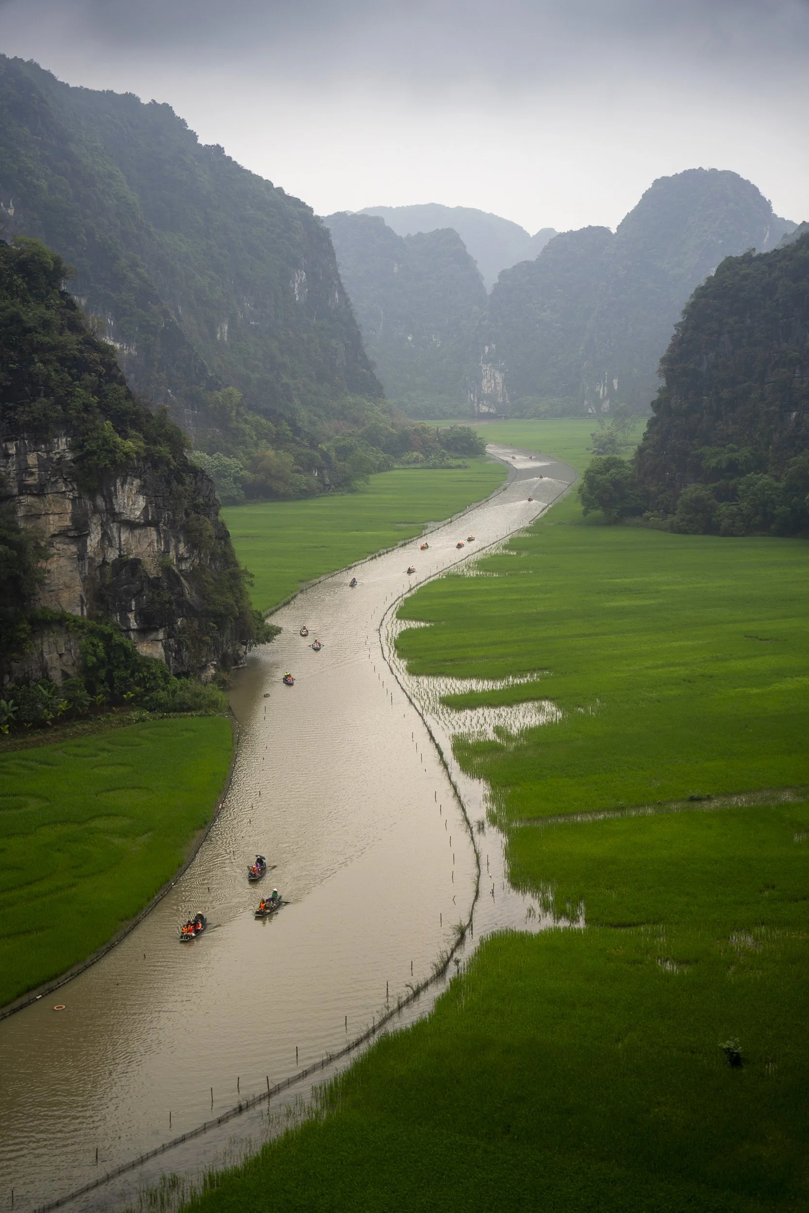 A winding river flowing through a lush green valley surrounded by steep, forested mountains on an overcast day.