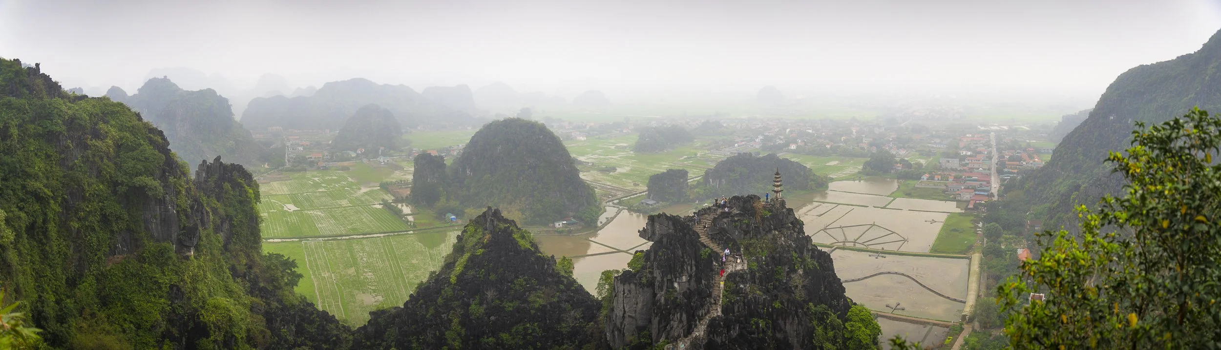A panoramic view of lush green mountains, rice paddies, and a small village with a pagoda on a rocky hilltop, shrouded in mist.