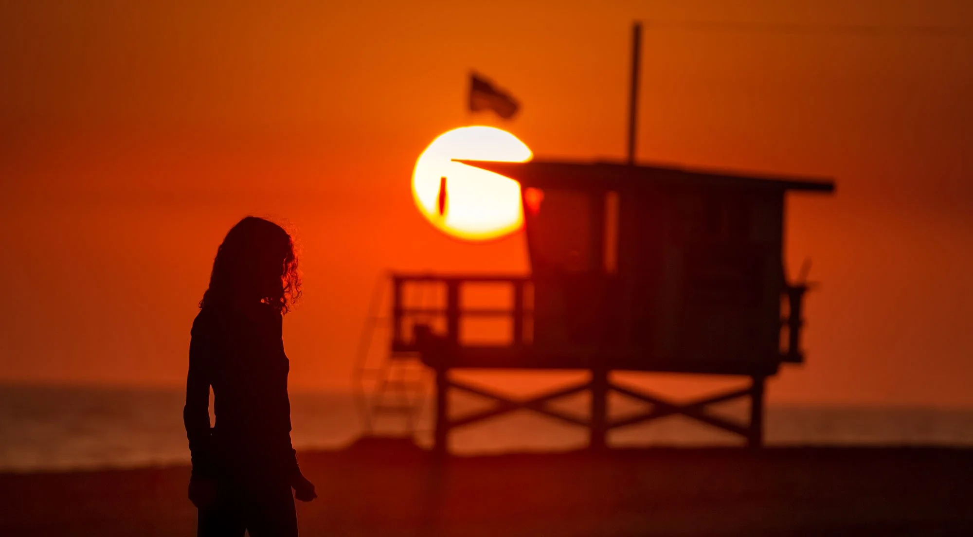 Silhouette of a woman with curly hair standing on a beach at sunset, with a lifeguard tower in the background and the sun setting over the ocean.
