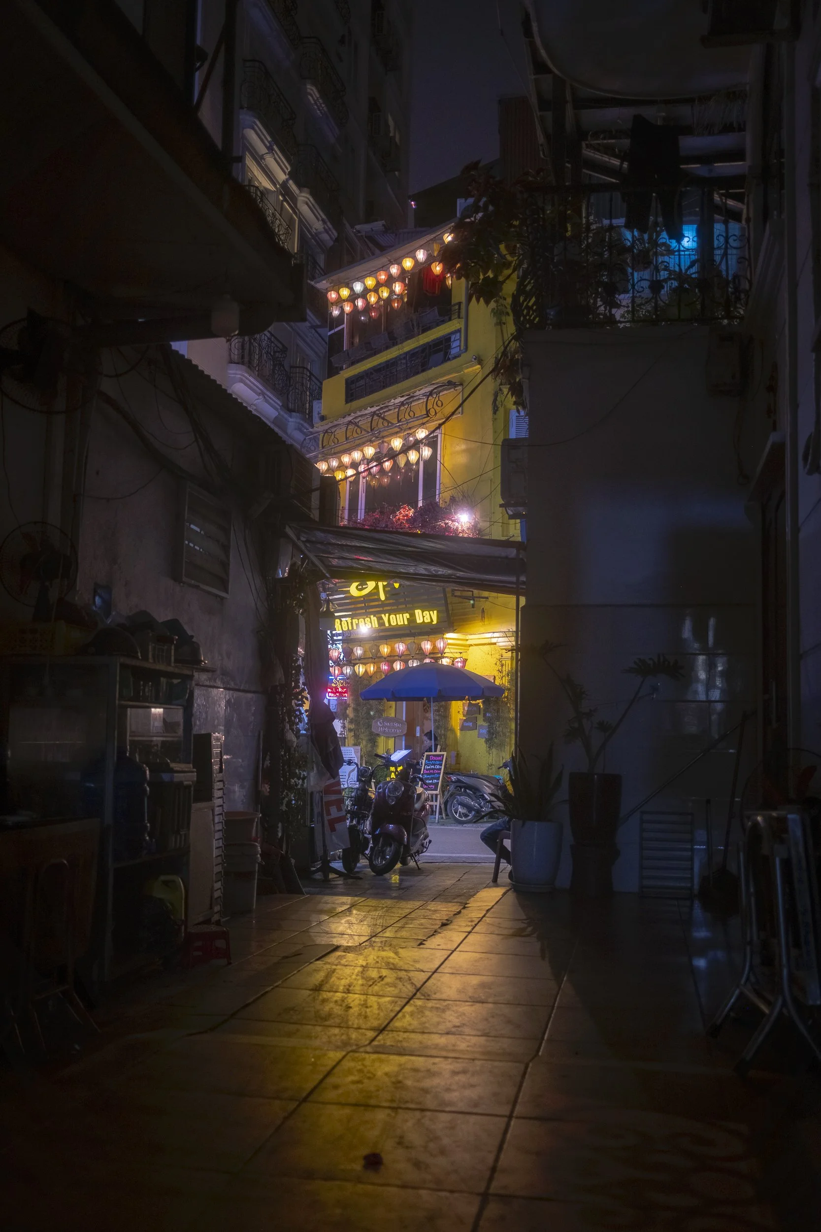 Nighttime view of a narrow street with illuminated lanterns, bikes, and a blue umbrella outside a building in a city, possibly in Vietnam.