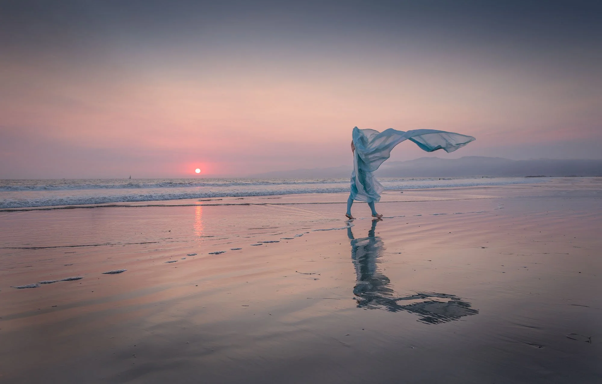 Person walking on the beach during sunset, holding a flowing white cloth, with the reflection visible on the wet sand.